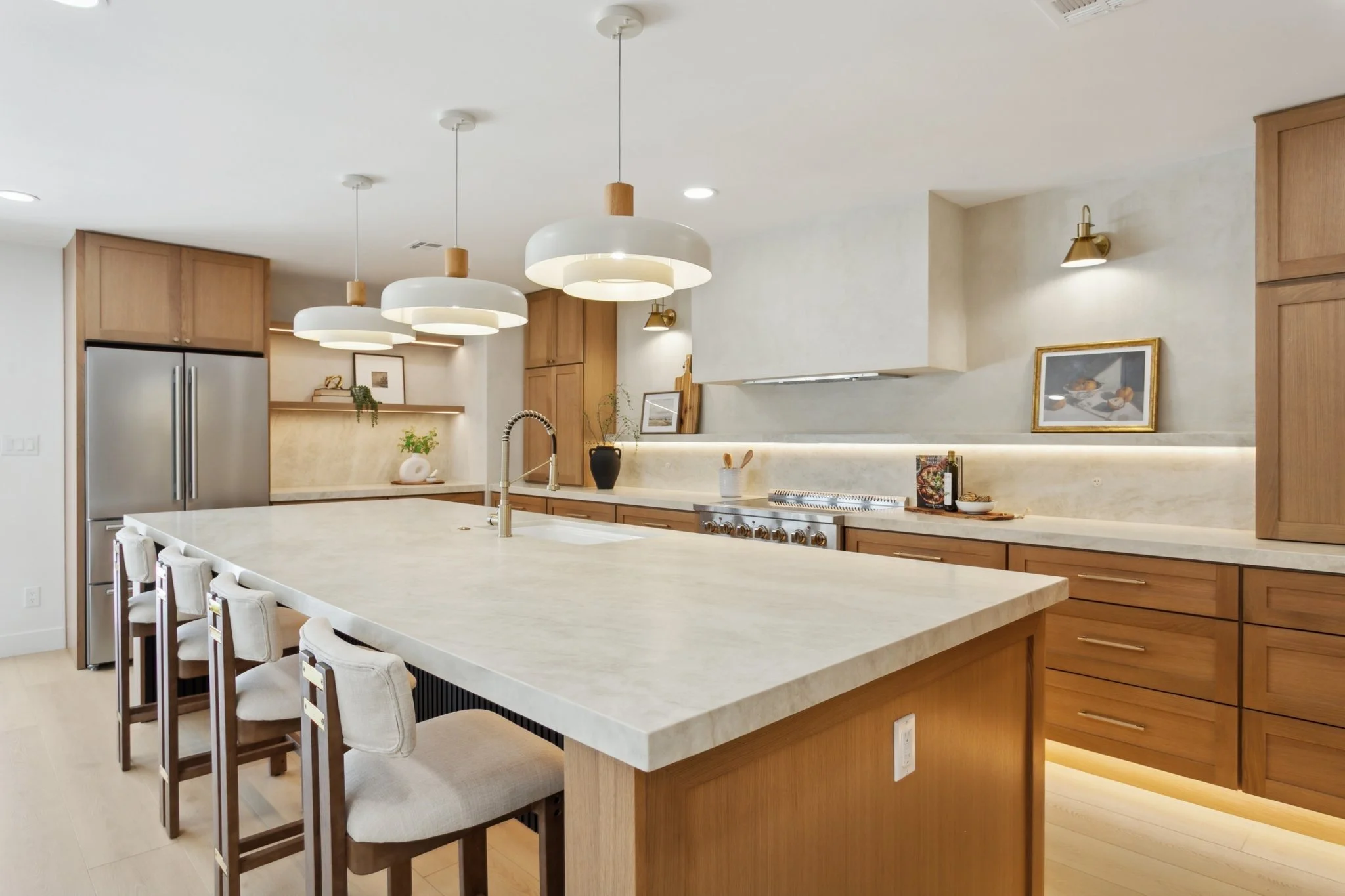 Modern kitchen with a large island, wooden cabinets, stainless steel refrigerator, and white pendant lights.