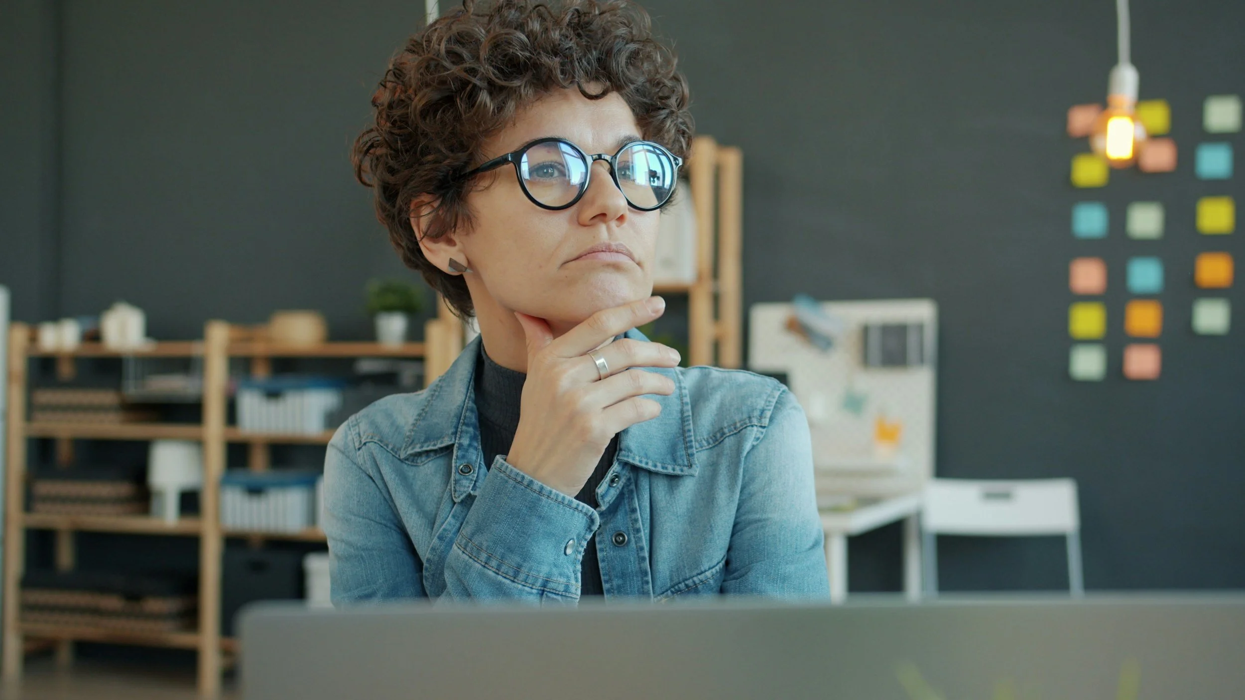 A woman with short, curly hair and glasses is looking thoughtfully at a computer screen, with her hand resting on her chin, in an office or creative workspace.