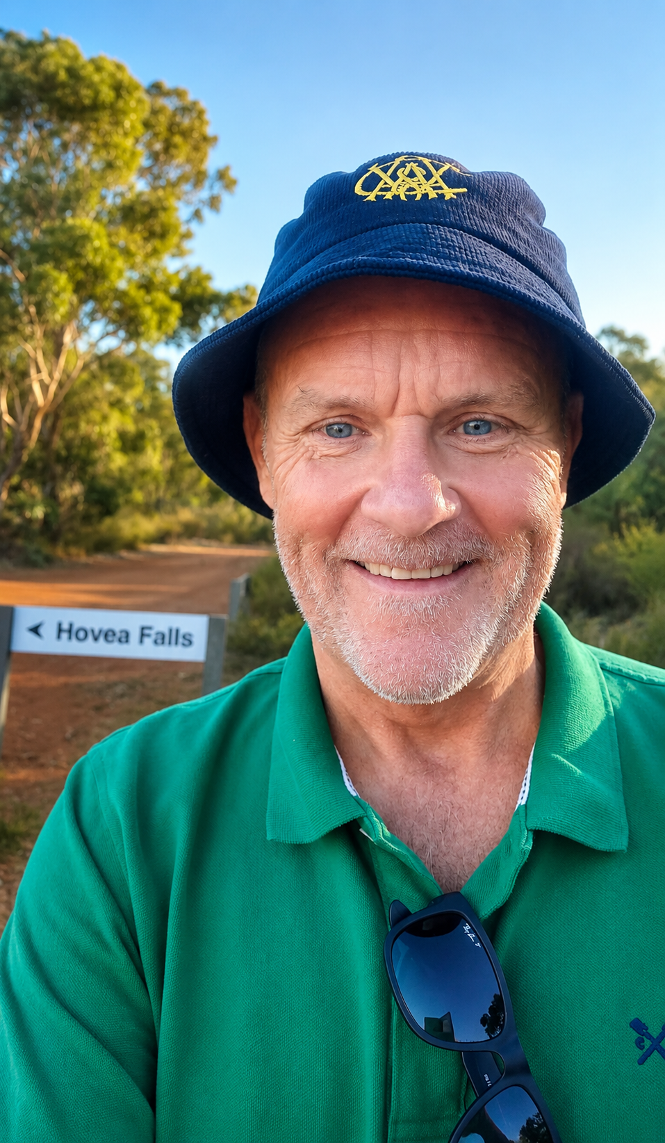 Smiling man with a beard wearing a blue bucket hat with yellow emblem, green shirt, and sunglasses hanging from shirt collar, outdoors in a sunny environment, with a sign that reads 'Hovea Falls' in the background.
