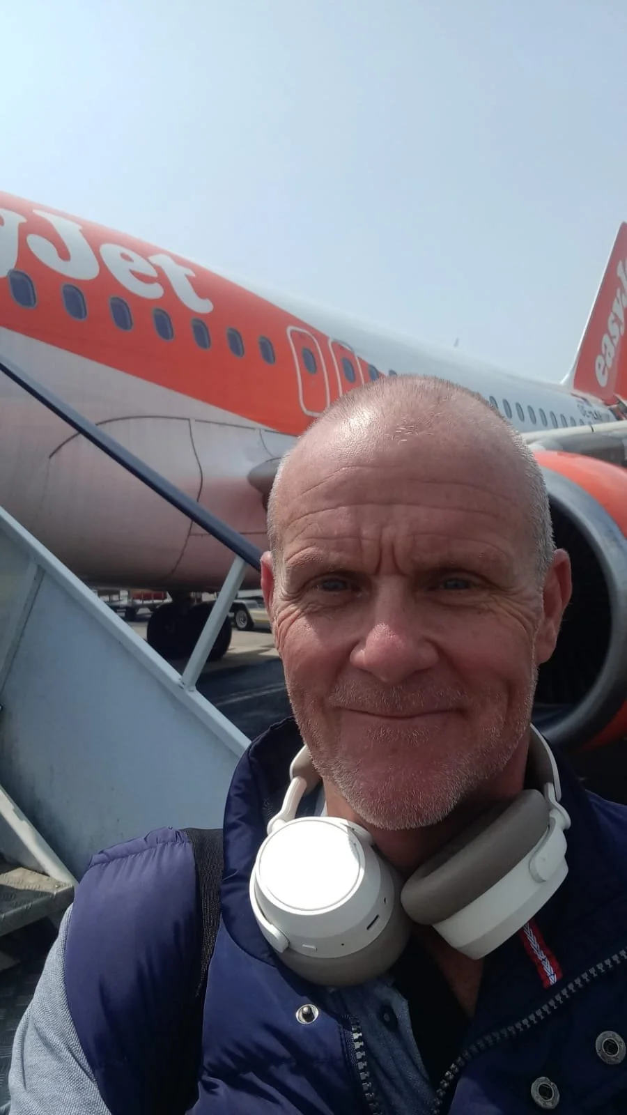 A man with a shaved head and beard taking a selfie at an airport, with an easyJet airplane in the background.