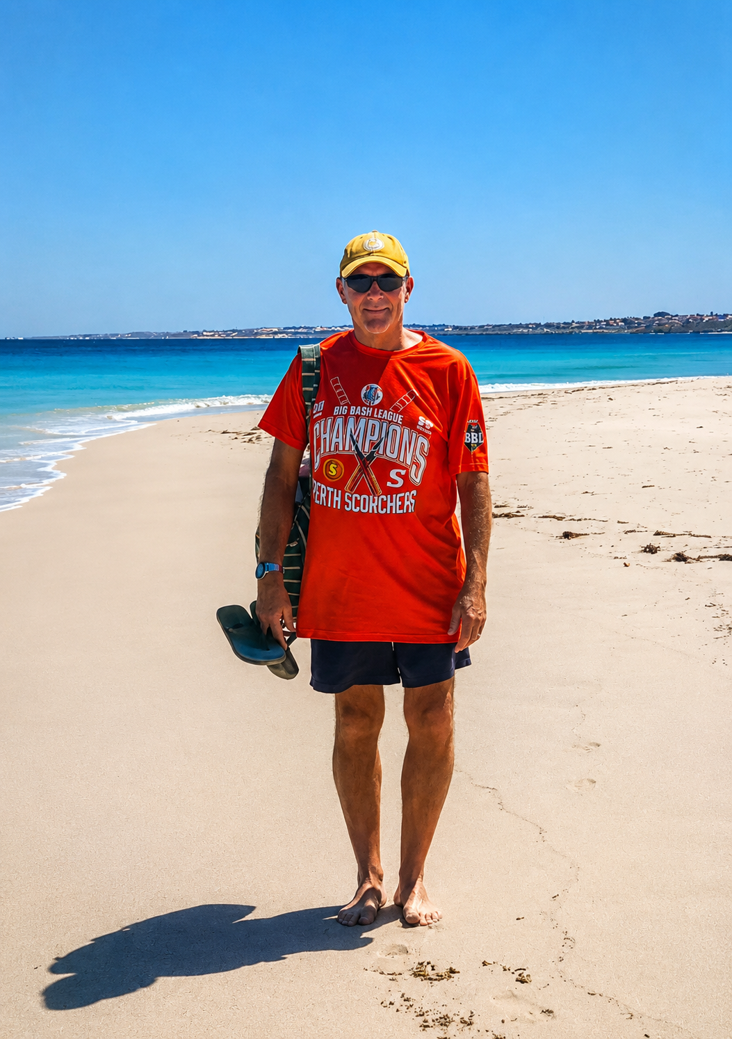 A man in a red sports jersey and shorts, wearing sunglasses, a yellow cap, and carrying flip flops, walking on a sandy beach with the ocean and a clear blue sky in the background.