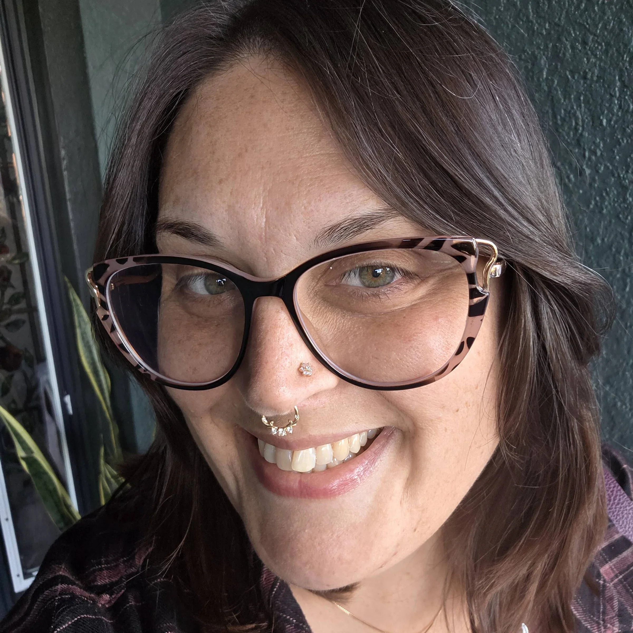 Close-up of a woman smiling, showing her teeth, wearing sunglasses with a tortoise shell pattern, with dark brown hair, a nose piercing with a small jewel, and a necklace. The background features a textured wall and part of a building or window.