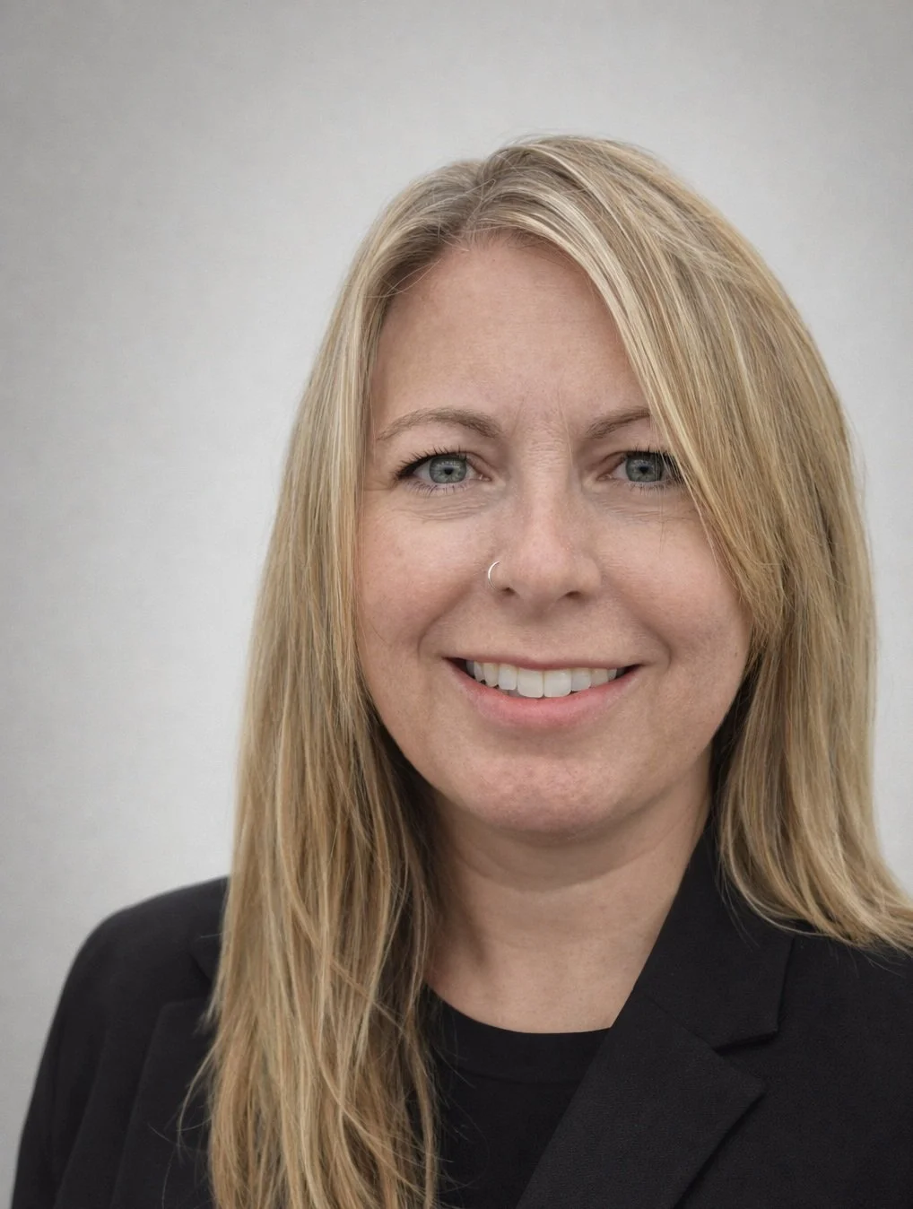 A woman with long blonde hair, blue eyes, and a nose ring, smiling, dressed in a black blazer, against a plain light-colored background.
