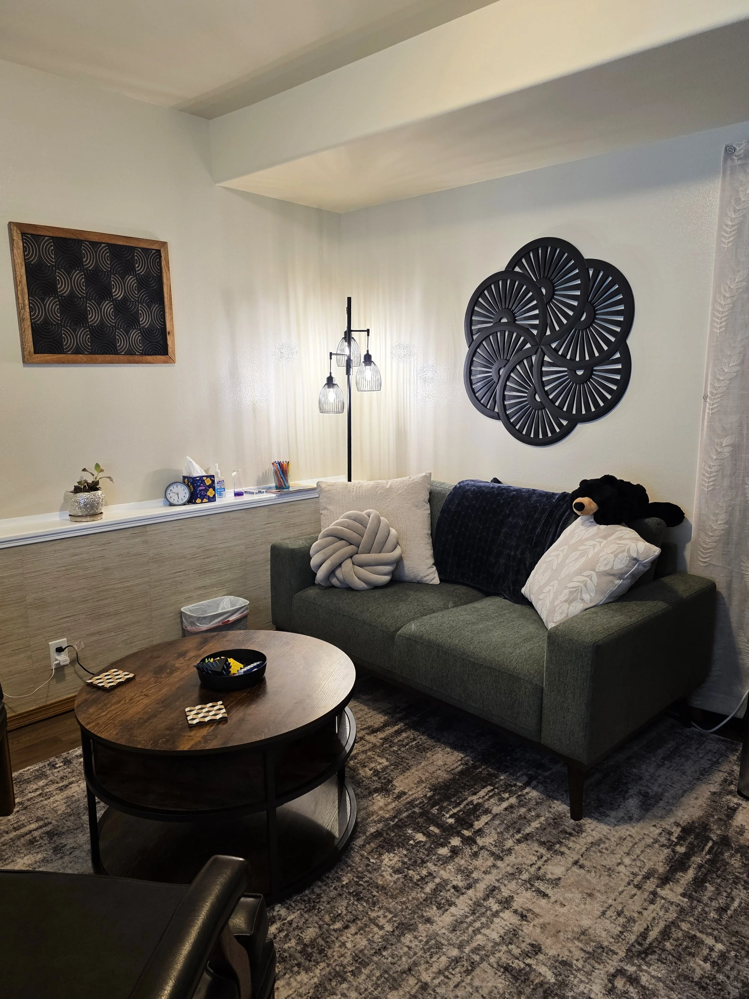 A cozy living room with a green couch, decorated pillows including one with a black bear plush toy, a dark oval coffee table with board games, a patterned rug, a white wall with black geometric wall art, a wall-mounted chalkboard, a corner lamp with three lights, a small potted plant, a clock, tissue box, and some writing utensils, and a sheer curtain on the right side.