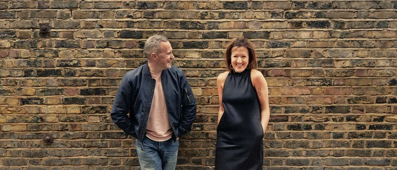 A man and a woman standing against a brick wall. Andy Jex and Larissa Vine. Both smiling and looking at each other. The man is wearing a navy jacket and jeans, and the woman is in a black sleeveless dress.