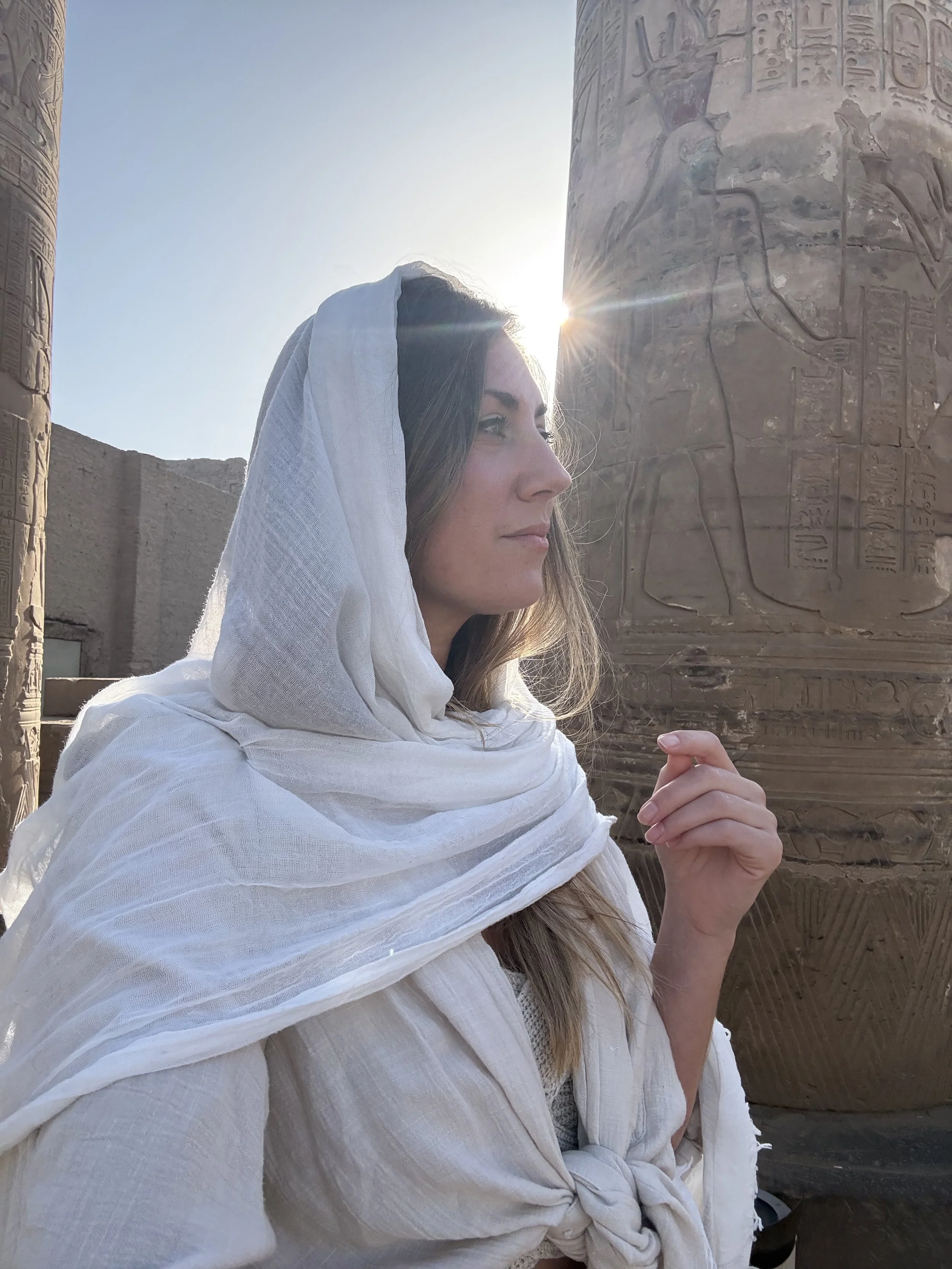 A woman dressed as an ancient Egyptian, wearing a white headdress and robes, standing next to carved hieroglyphic stone pillars looking ahead.