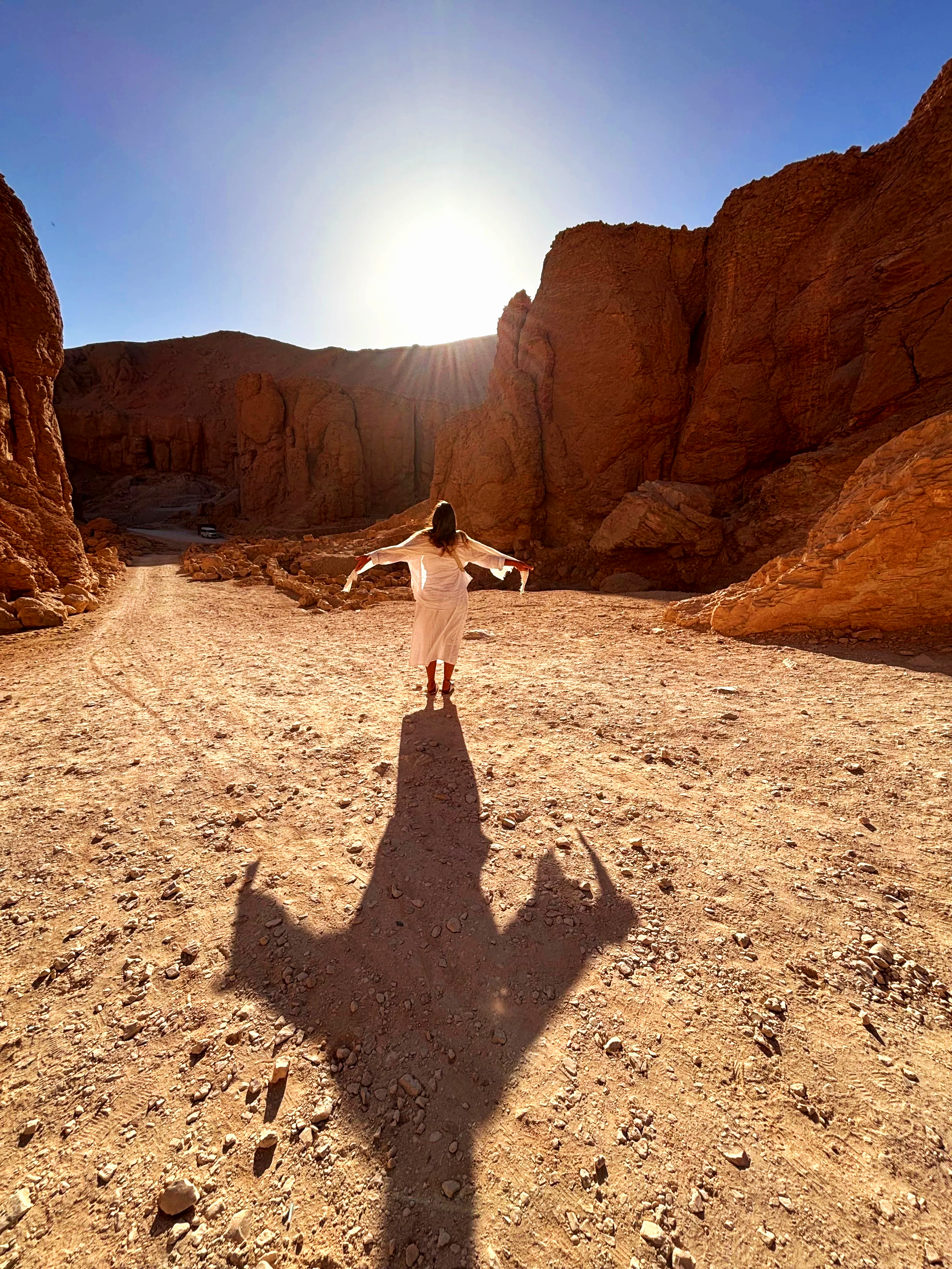 A woman standing on a rock in a desert canyon with large reddish-orange rock formations, during sunset with the sun low on the horizon casting her long shadow.