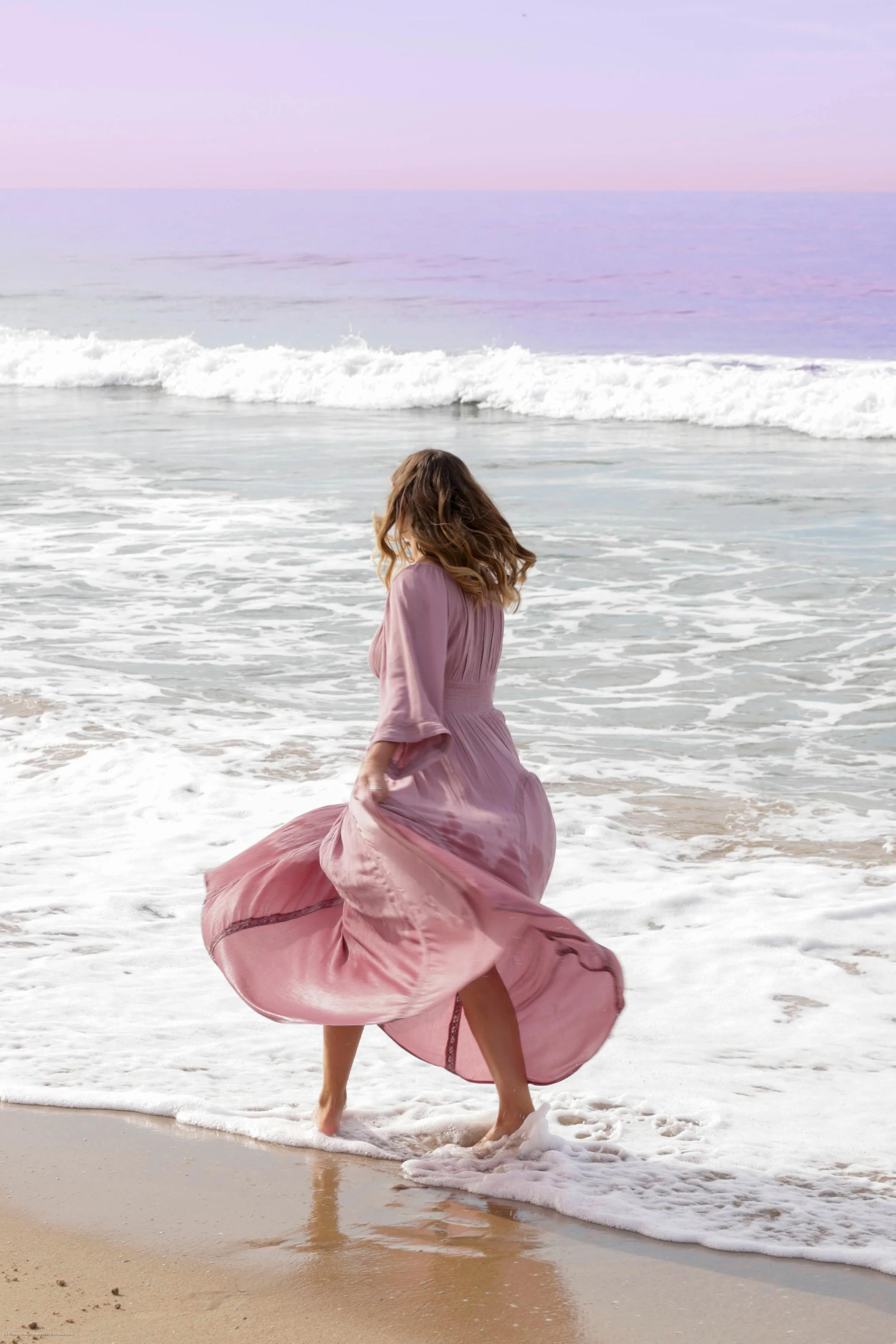 A woman in a flowing pink dress standing in the shallow surf at the beach, with waves approaching and the horizon visible in the background.