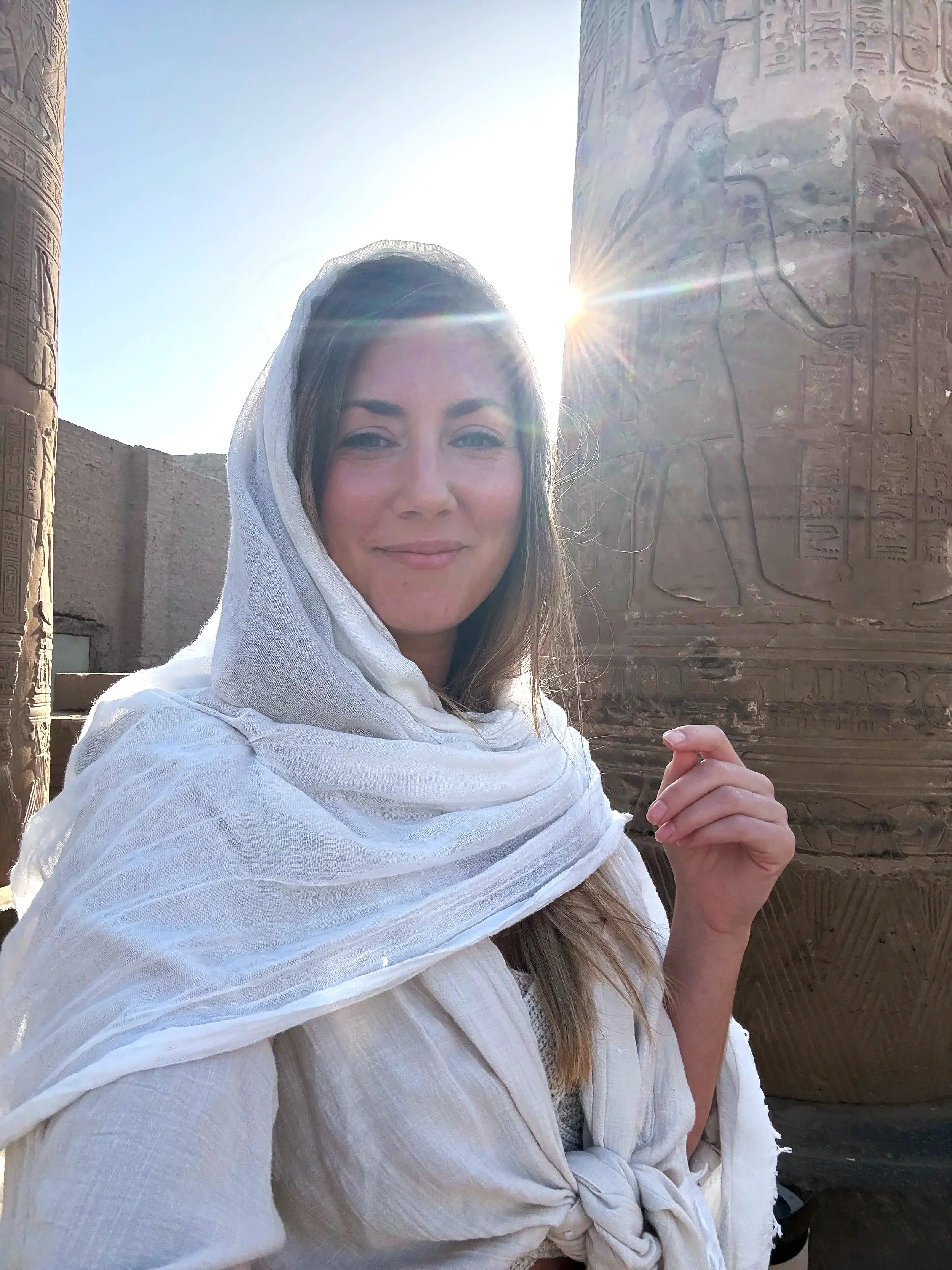 A woman dressed in ancient Egyptian costume standing in front of hieroglyphic carvings on stone columns, with sunlight shining in the background.