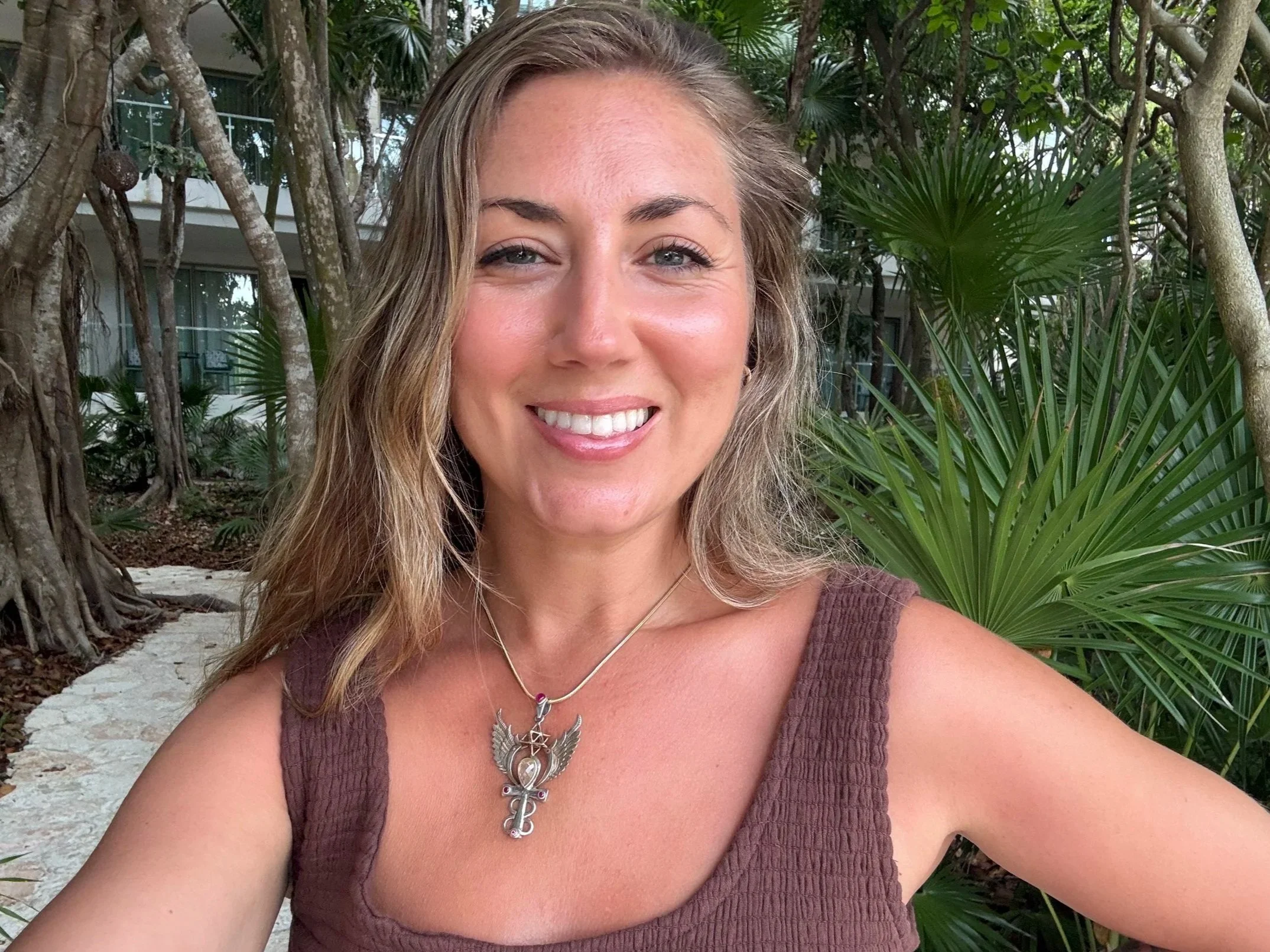 Close-up of a smiling woman with long wavy blonde hair, wearing a silver necklace with dangling charms, a brown sleeveless top, outdoors among tropical plants and trees.