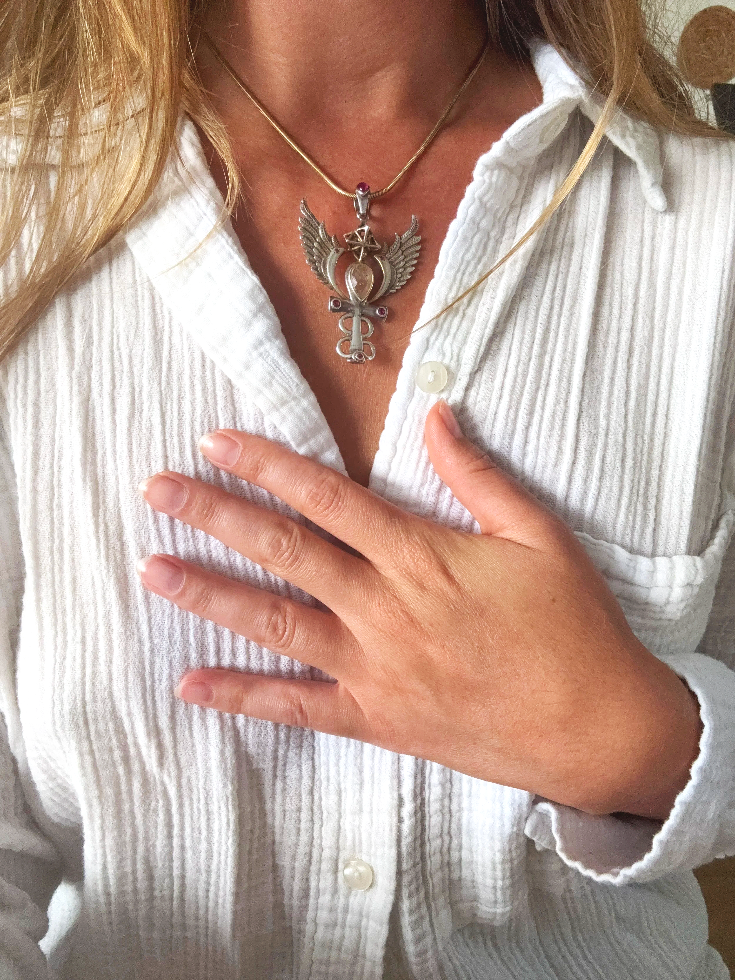 Close-up of a woman wearing a white, textured shirt with an open collar, a silver necklace with a detailed winged pendant, and a hand with manicured nails resting on her chest.