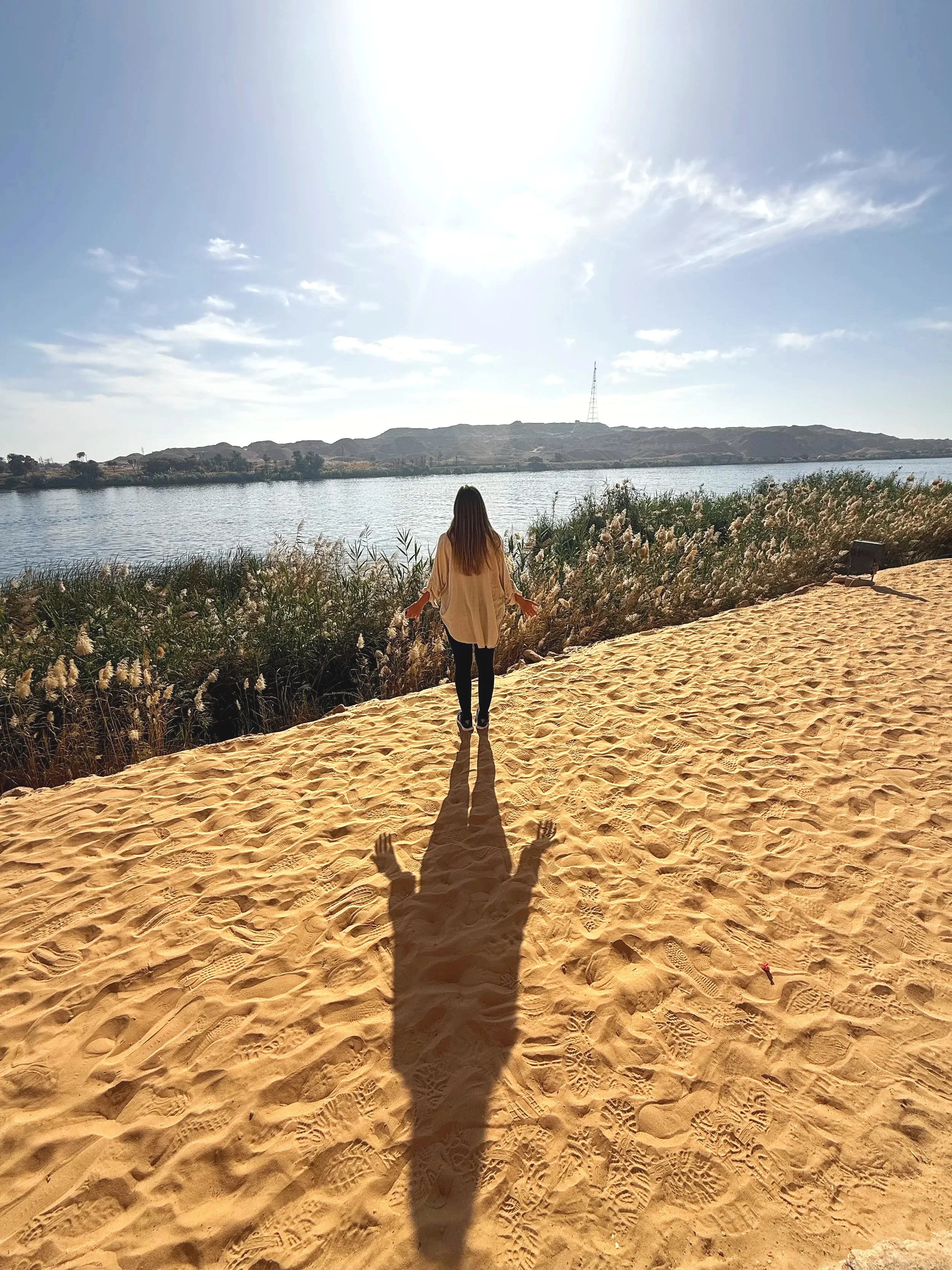 A person with long hair wearing a beige top and black pants standing on sandy ground near a body of water, with hills and a radio tower in the background, during a sunny day.