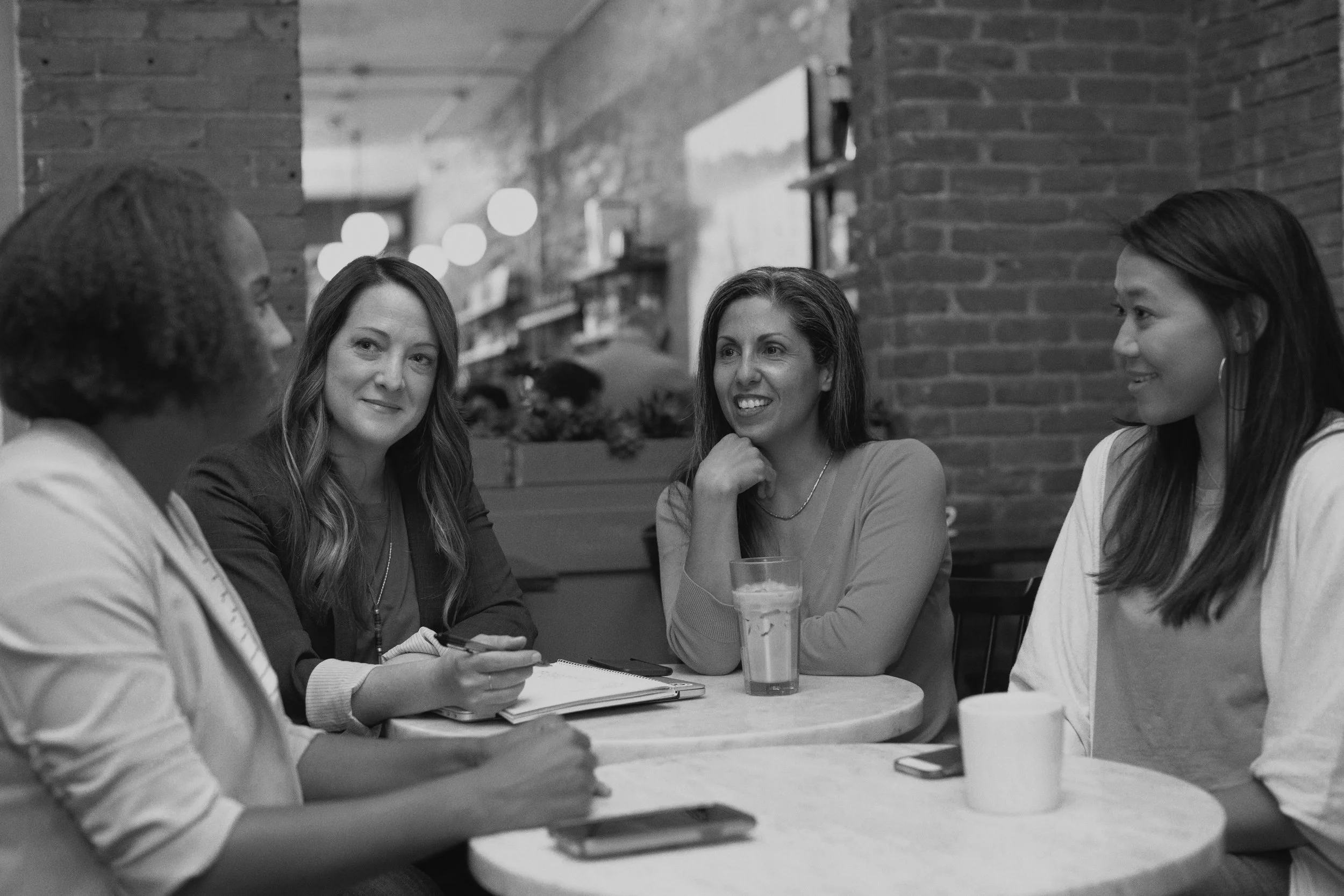 A group of five women sitting around a table in a cafe or restaurant, engaged in conversation. The scene is in black and white with a brick wall background, and drinks are on the table.