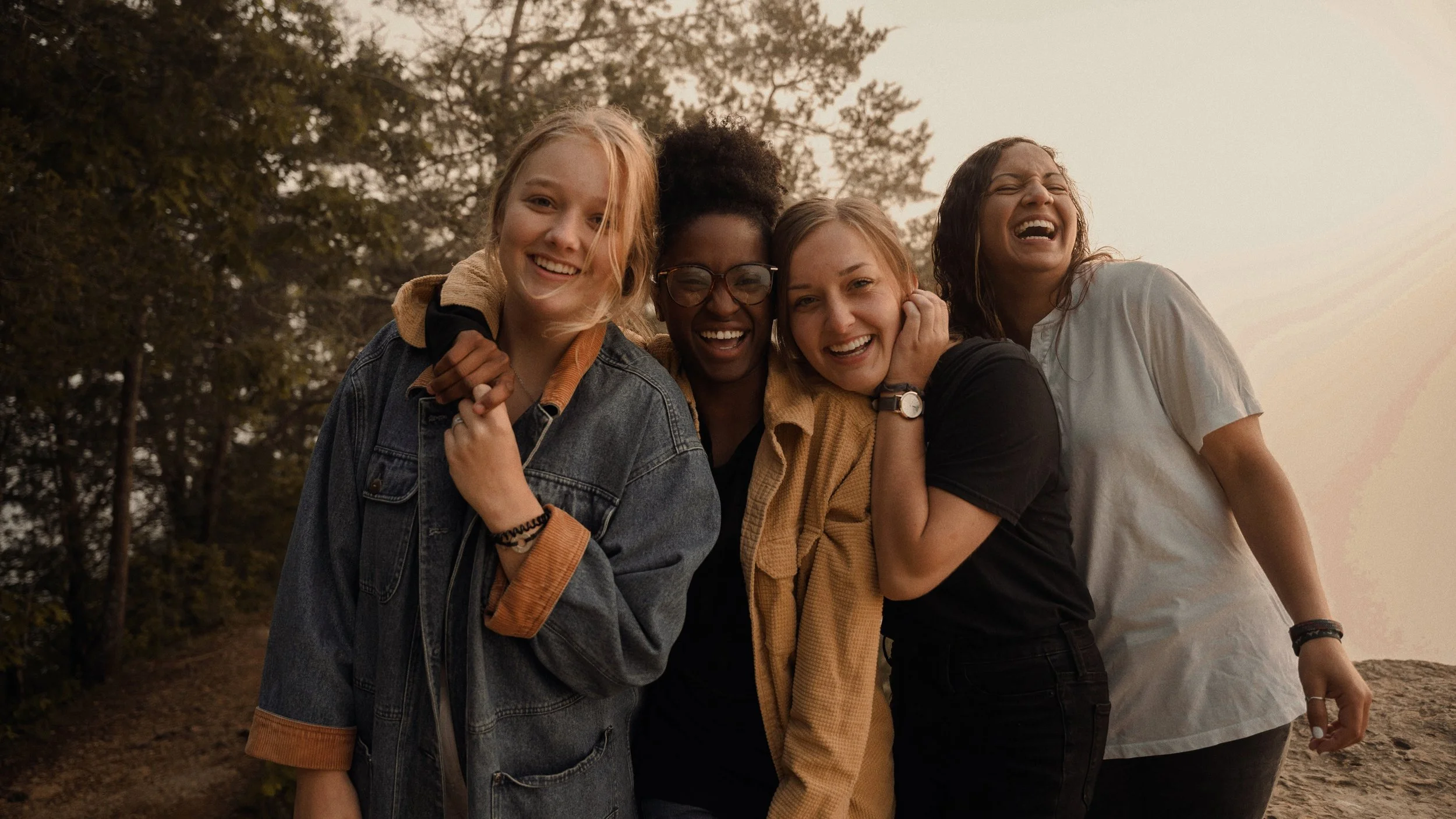 Four women are outside in a natural setting, smiling and laughing together. They are standing close with their arms around each other, enjoying each other's company.