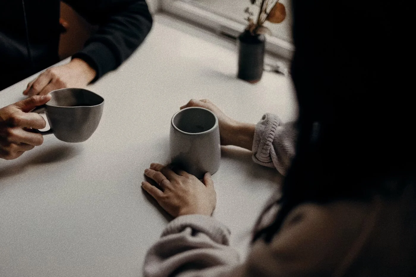 Two people exchanging coffee mugs at a white table, with one person's hands resting on the table. The image is taken from above, and the scene appears to be in a cozy indoor setting.