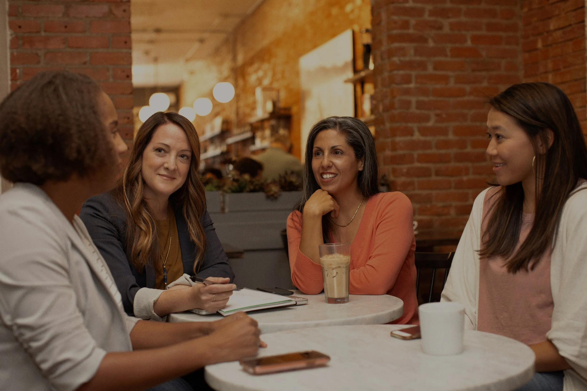 Four women sit around a table, engaged in conversation in a cozy, brick-walled cafe. One woman appears to be listening while others talk, with drinks and notebooks on the table.