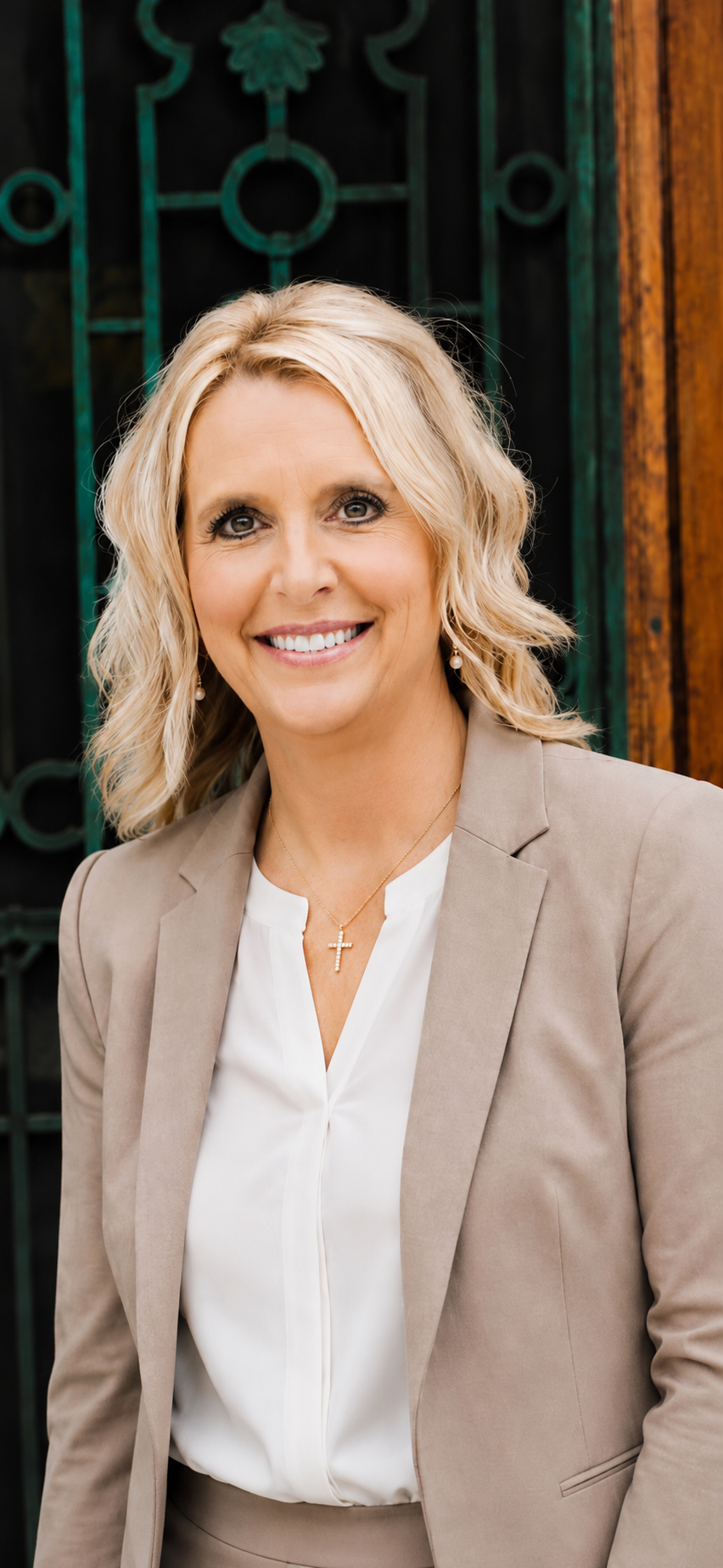 A woman with blonde curly hair wearing a beige blazer and white blouse, smiling at the camera, standing outdoors with a decorative green iron gate and wooden wall behind her.