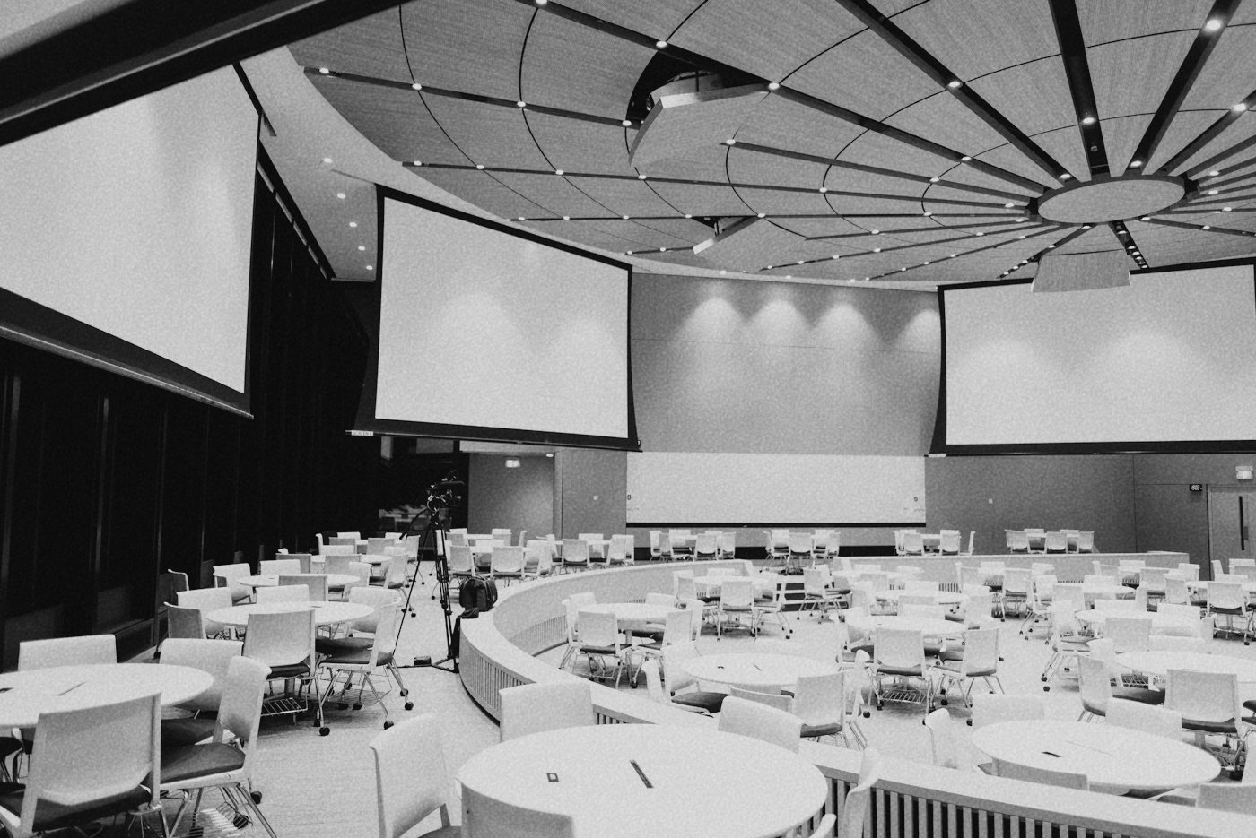 An empty conference room with round tables and chairs, large projection screens, and a camera on a tripod, ready for an event.