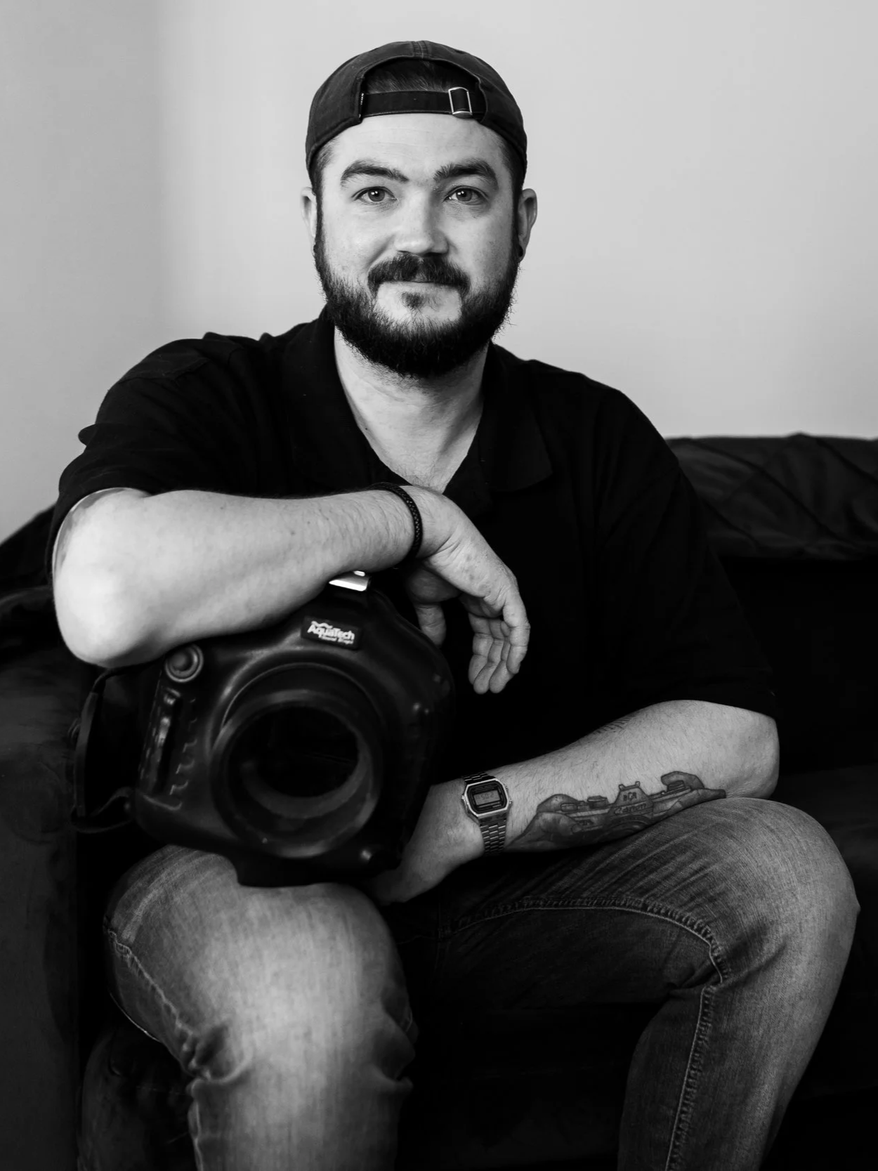 Black and white photo of a man with a beard wearing a backwards cap, sitting on a couch, holding a camera, with a tattoo on his left arm, a watch, and a bracelet.