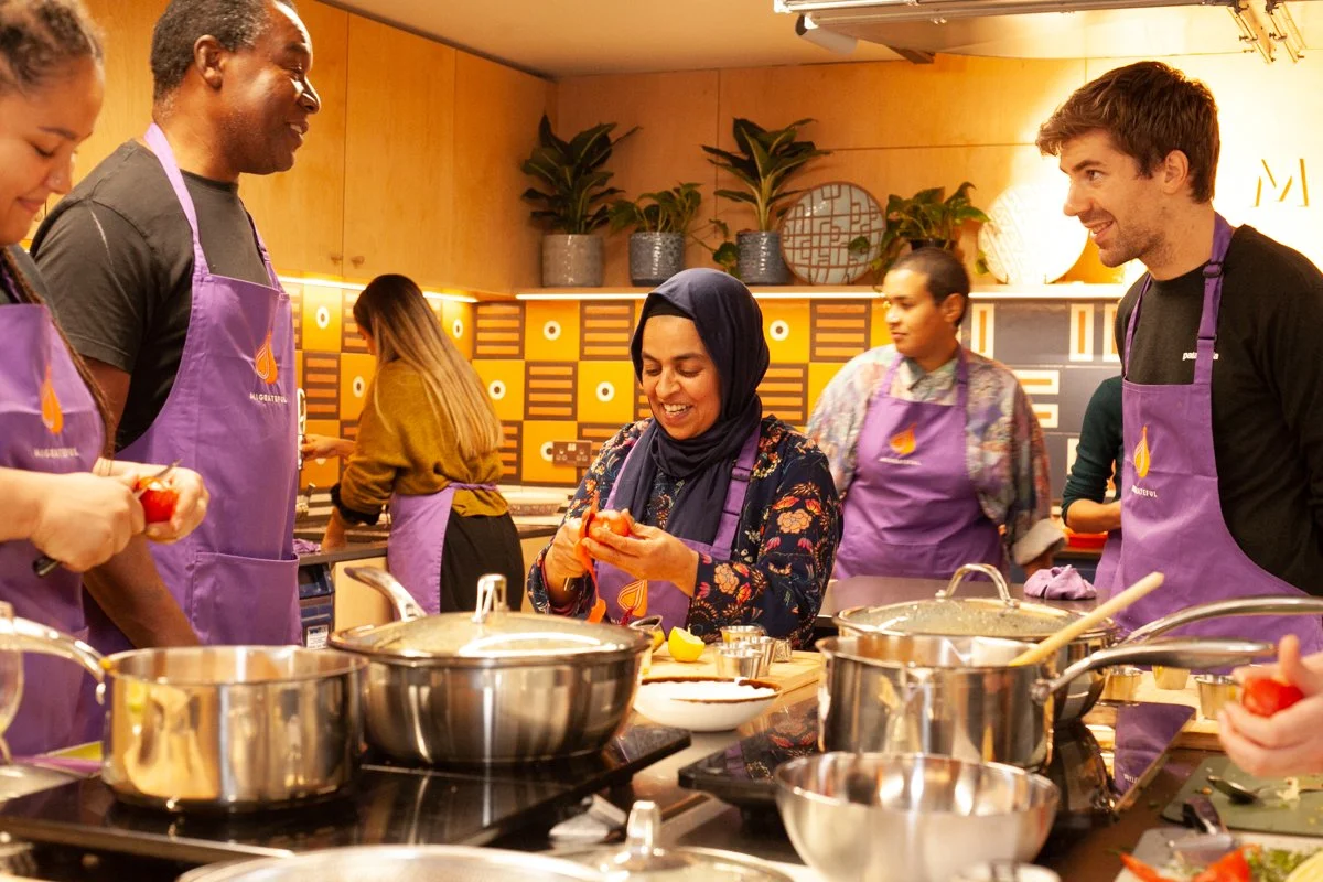 People participating in a cooking class, wearing purple aprons, gathering around a kitchen counter, peeling tomatoes, with pots and cooking utensils on the counter, and a warm, modern kitchen background.