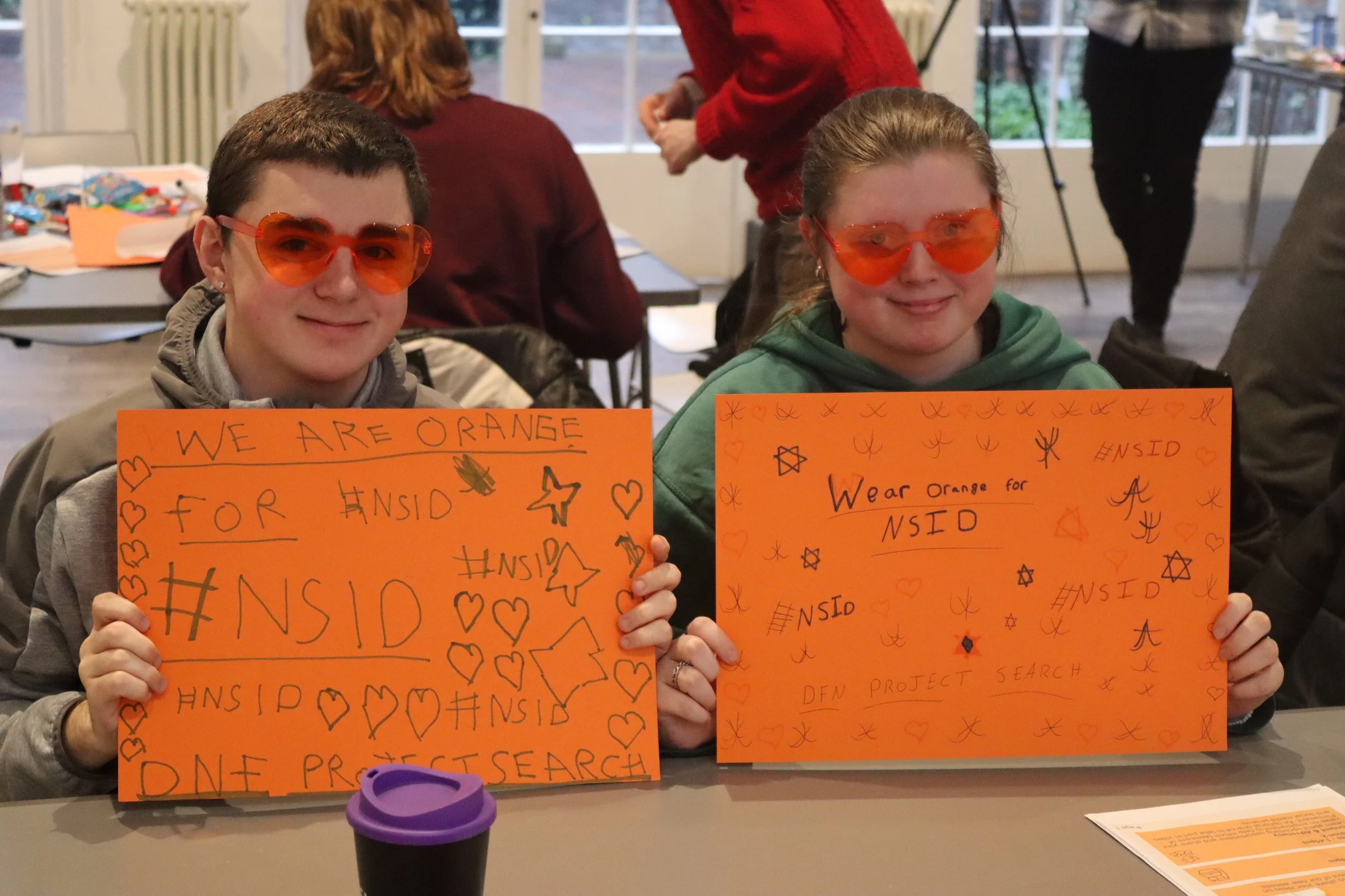 Two smiling people, one on each side, wearing orange sunglasses and holding orange signs with handwritten text and doodles, participating in a demonstration or event at an indoor location with tables, papers, and people in the background.