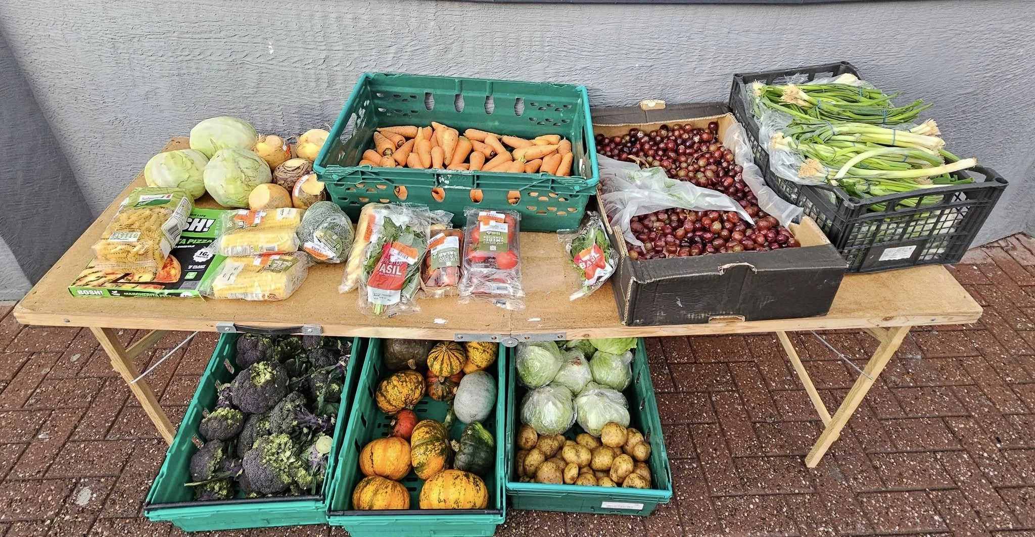 A table with various fresh vegetables and produce including cabbages, carrots, onions, broccoli, pumpkins, potatoes, and leeks, arranged in plastic crates and on the table surface.
