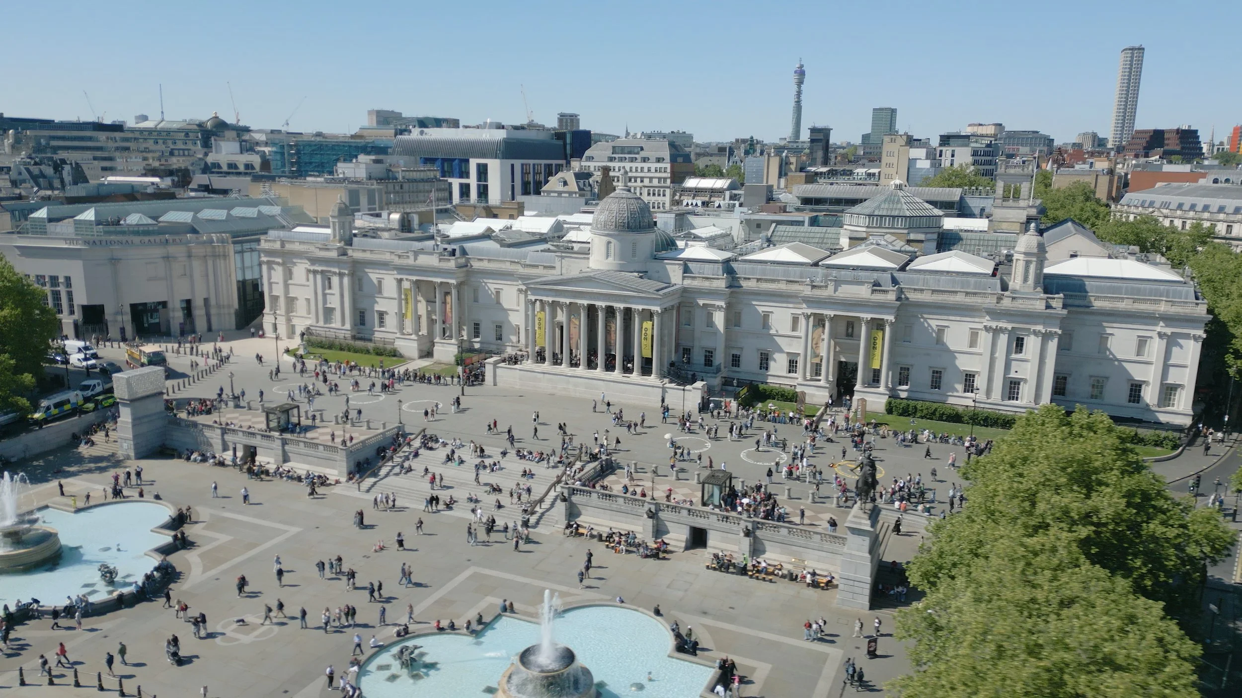 View of Trafalgar Square in London with the National Gallery building and many people walking and sitting around the fountains.