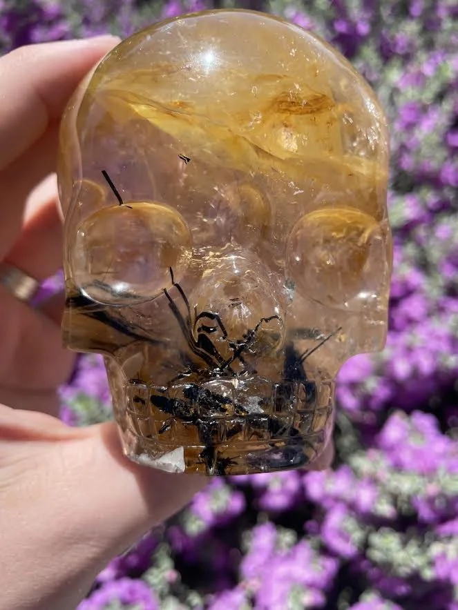 A clear skull-shaped gemstone with black inclusions, held up against a background of purple flowers.