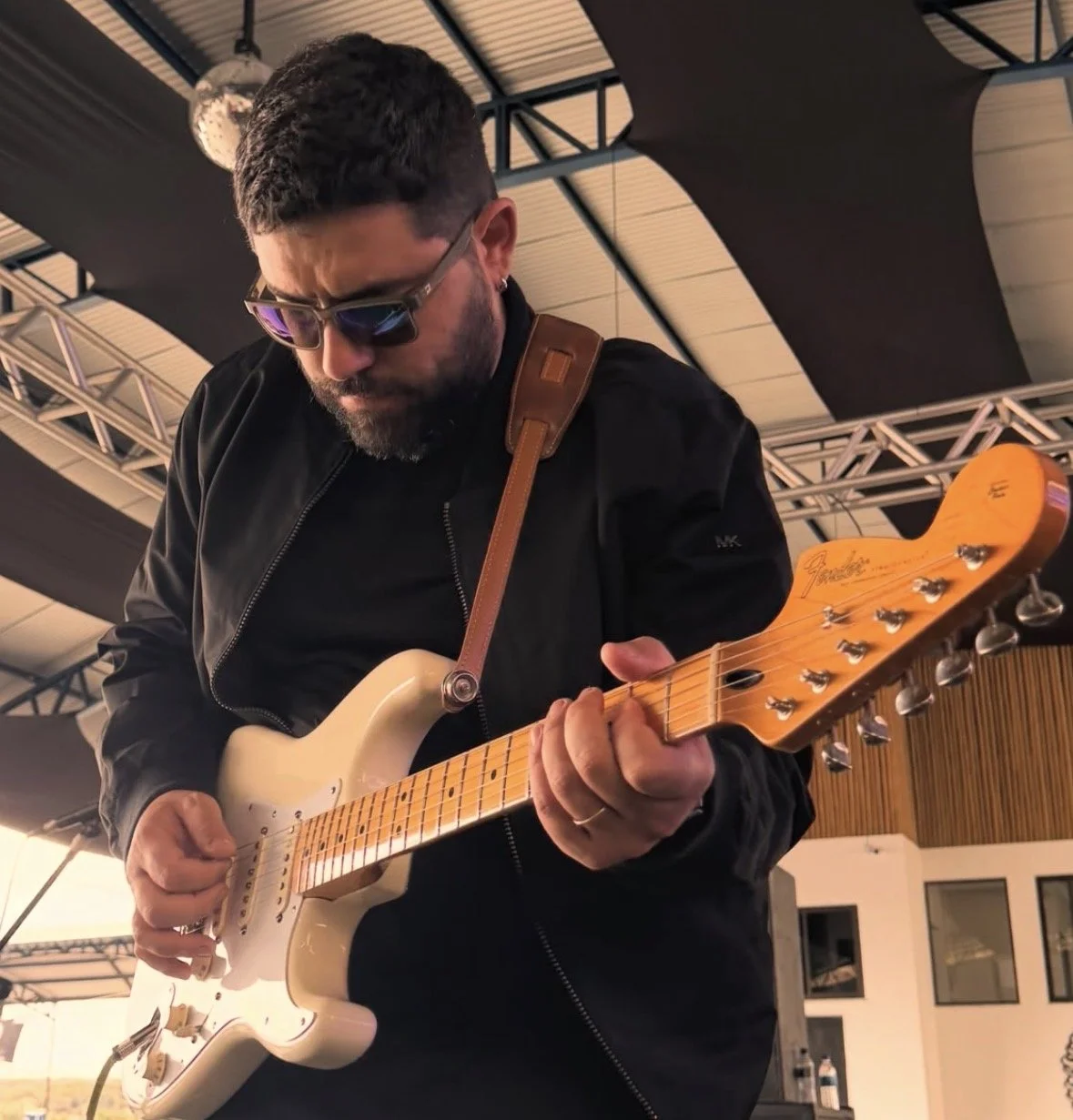 A man wearing sunglasses and a black jacket playing a Fender electric guitar on a stage.