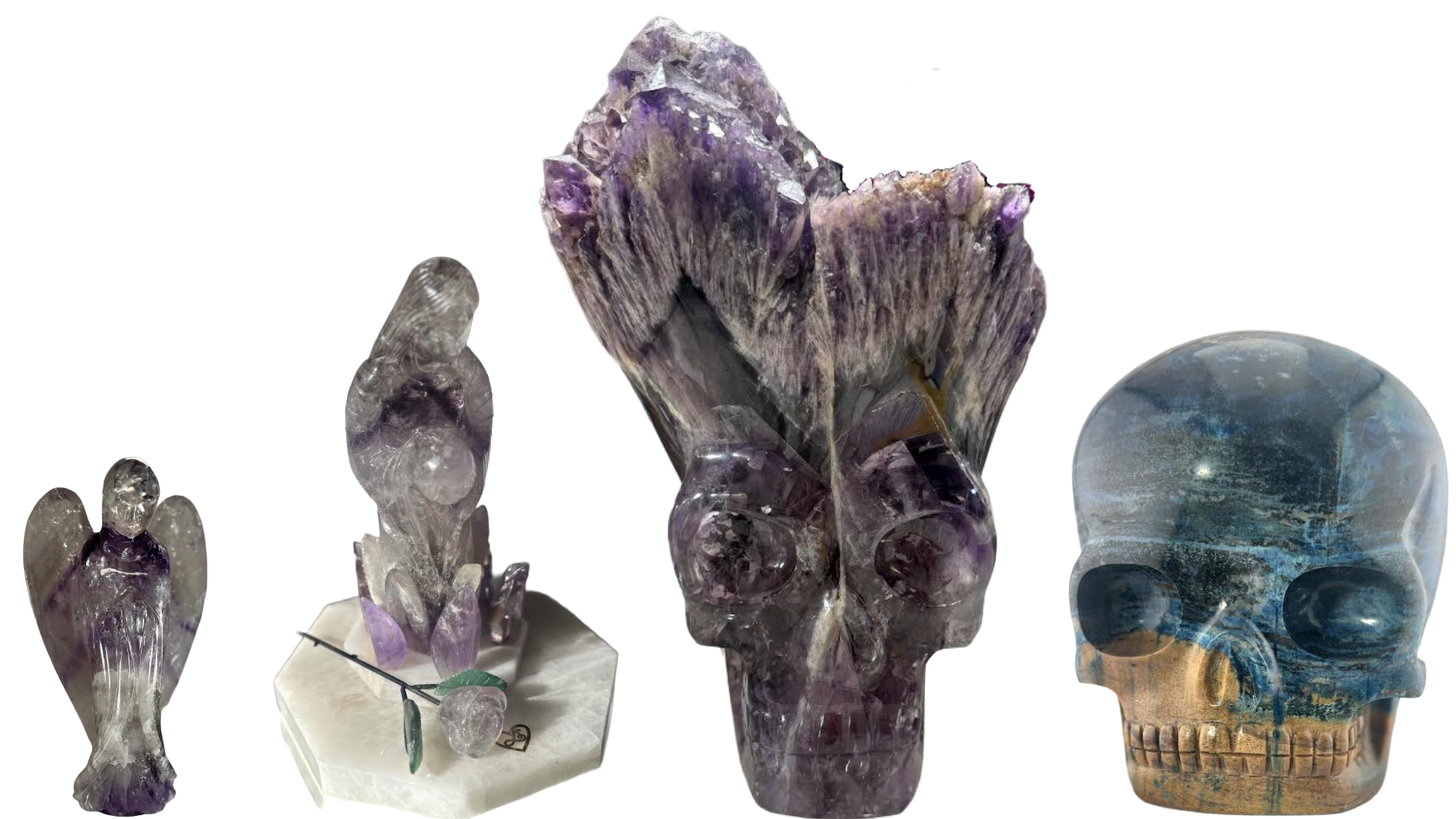 Collection of carved crystal and stone sculptures, including an amethyst skull, angel figure, and smaller figurines, displayed on a white surface.