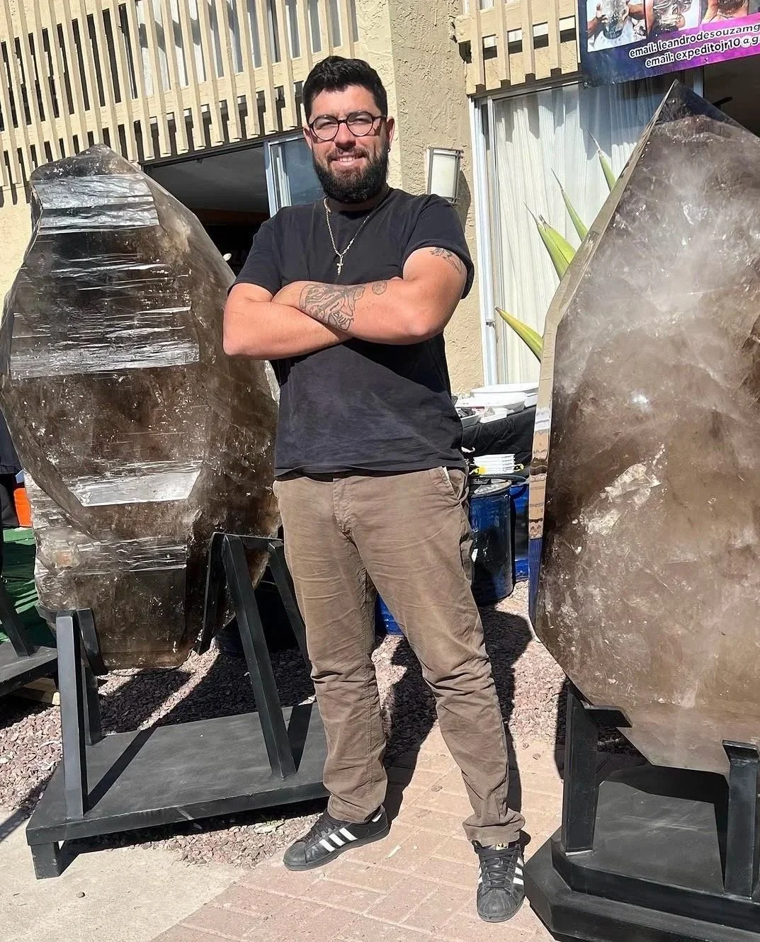 A man with glasses, a beard, and tattoos, standing outdoors with his arms crossed, posing next to two large, polished crystal sculptures.
