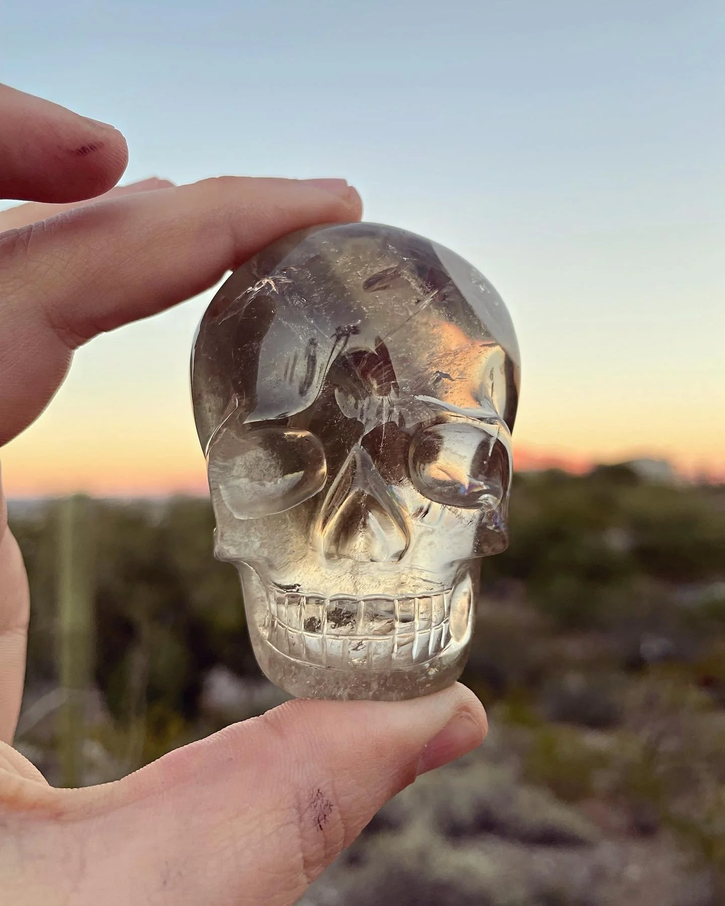 Hand holding a Garden Quartz Shaman Palm Skull against a sunset sky with horizon and landscape