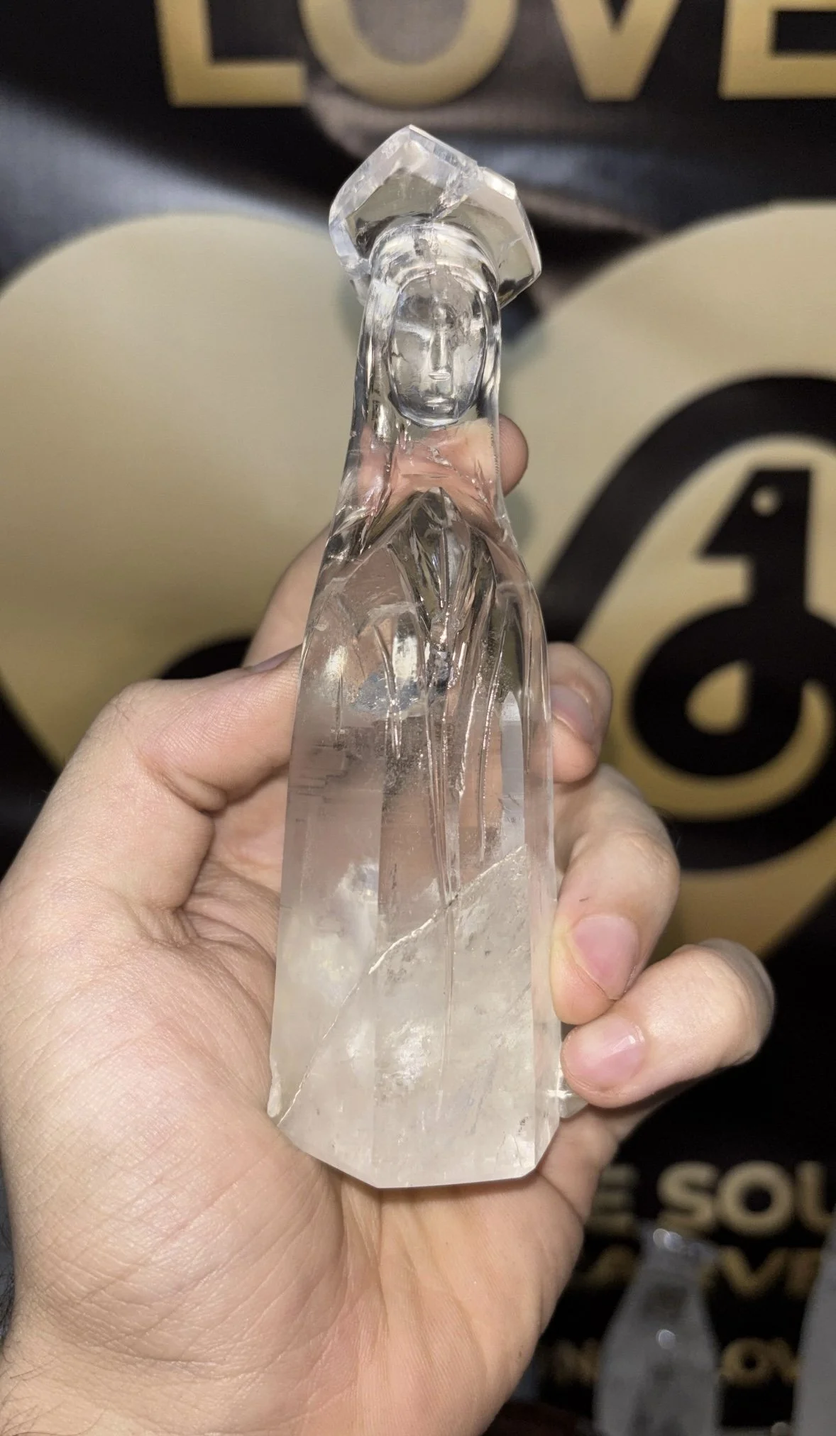 Close-up of a hand holding a carved clear quartz crystal point with a carved face detail near the top, with a background featuring part of a black display with gold text.