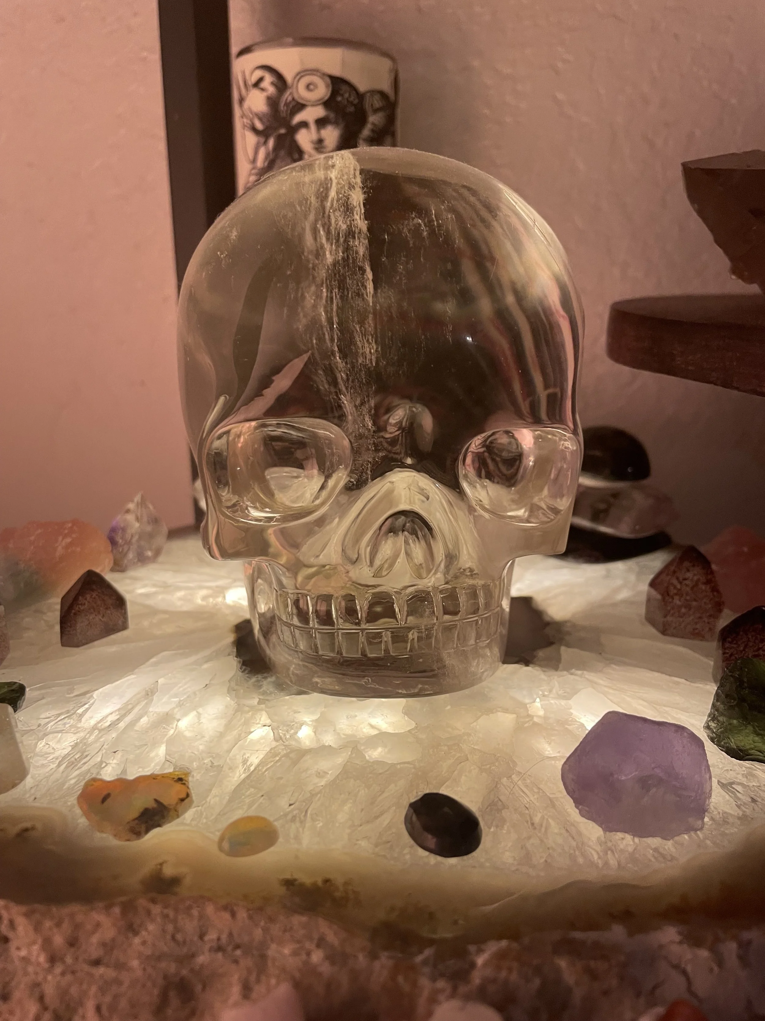 A glass skull on a lighted surface, surrounded by various colorful stones. In the background, there's a tall cup with a tribal face design on it.