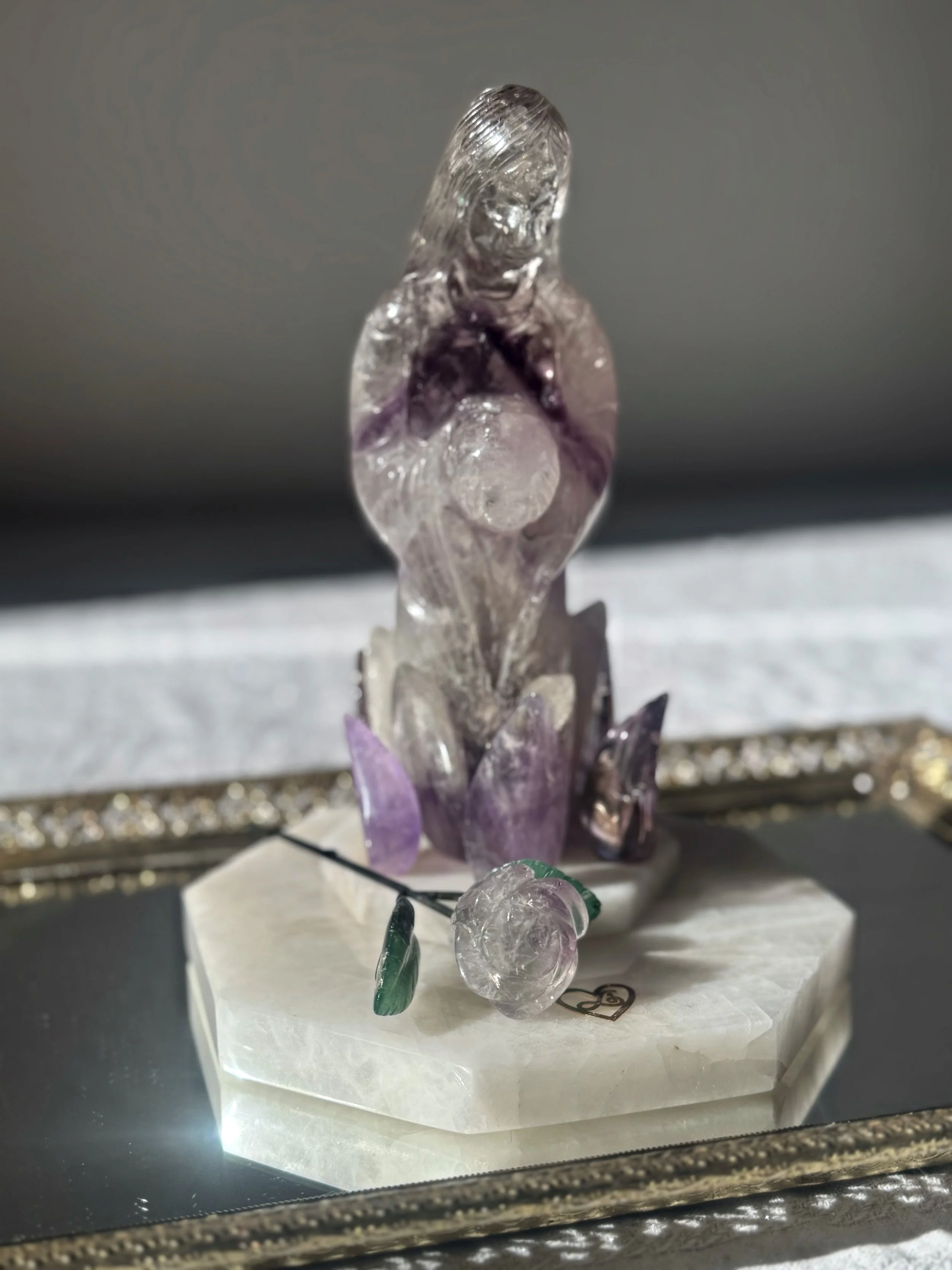 A small sculpture of a person sitting with their knees to their chest, made from clear and purple glass or crystal, displayed on a white base inside a glass case.