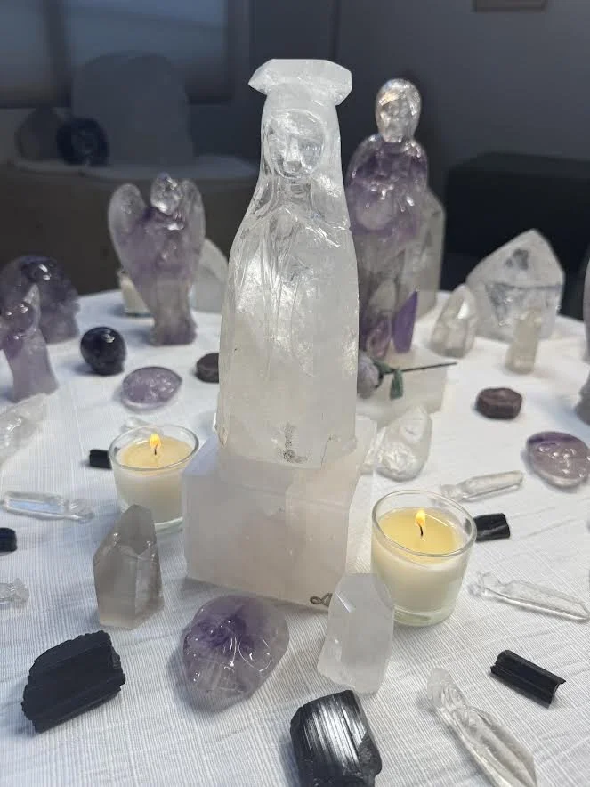 Crystals and gemstones on a table with two lit candles in glass holders, with a large Mother Mary Cathedral Quartz centerpiece carved by Leandro de Souza in the middle.