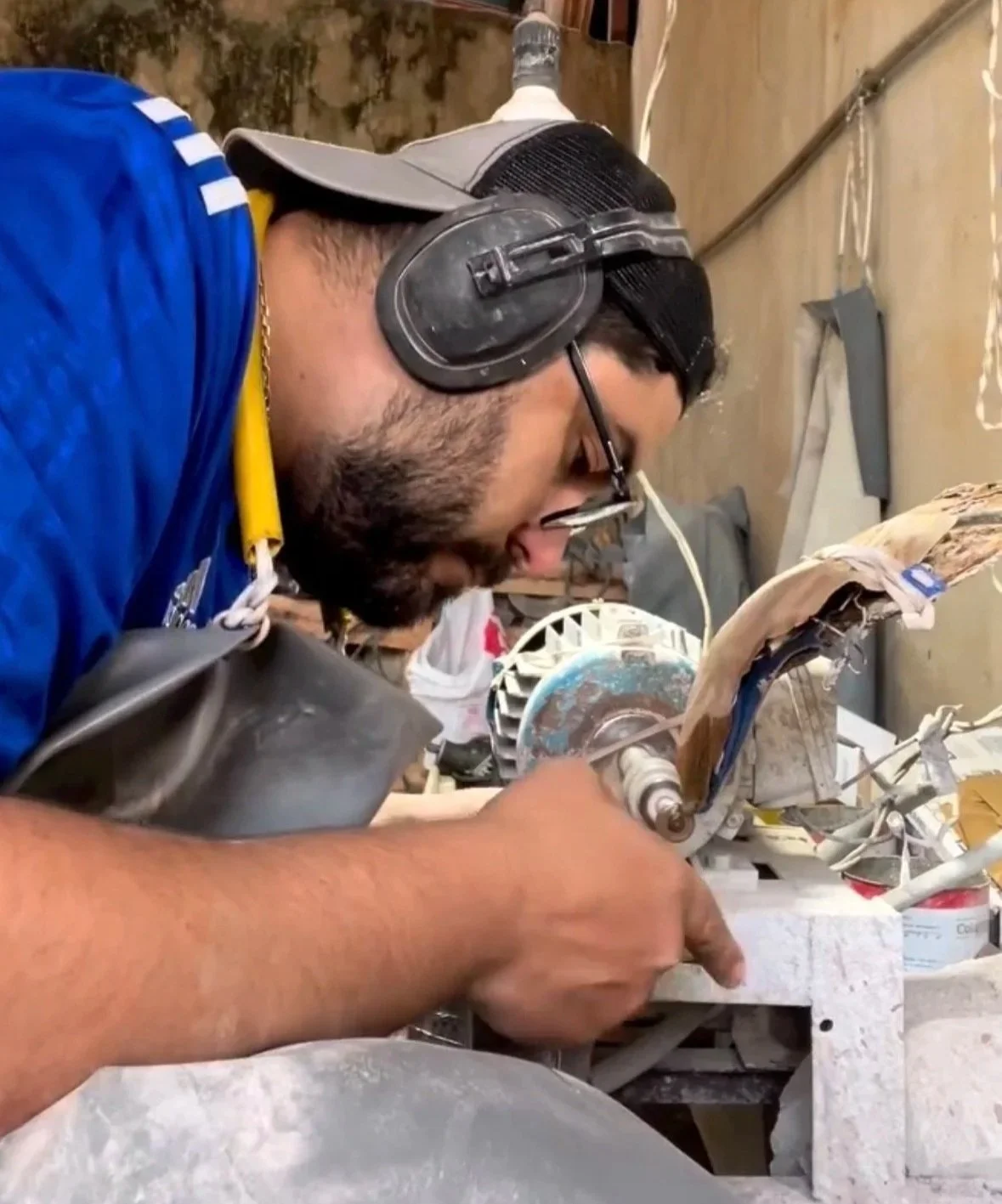 A man wearing safety headphones, glasses, and a cap is using a handheld power tool to work on a wooden object in a workshop.