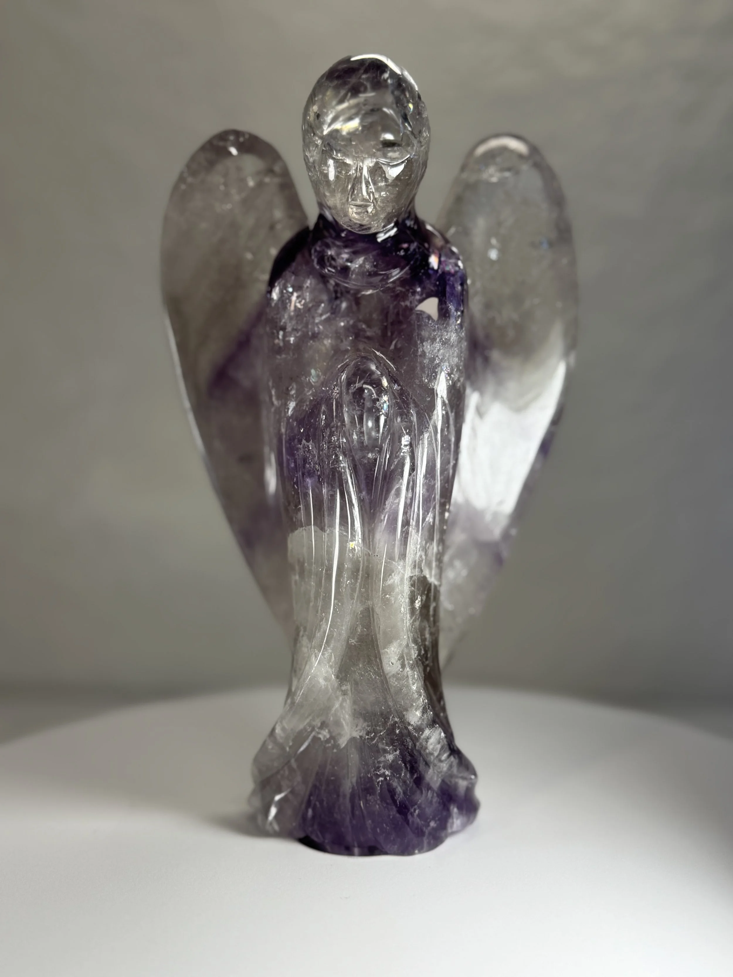 A small glass angel figurine with a clear face and purple body, standing on a white surface.