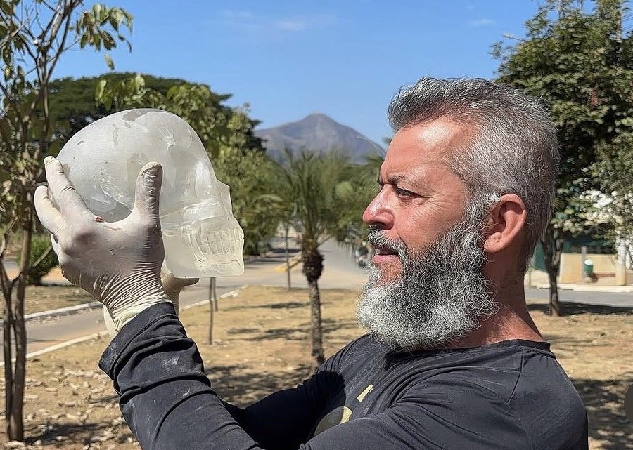 A man with a beard wearing a black long sleeve shirt holds a transparent skull in his gloved hand, outdoors with trees and mountains in the background.