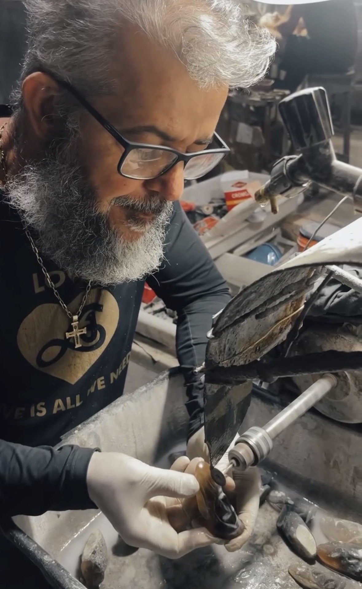 A man with glasses and a gray beard working on a motorcycle in a workshop, holding an old, worn motorcycle part in his hands, with tools and workshop items visible in the background.