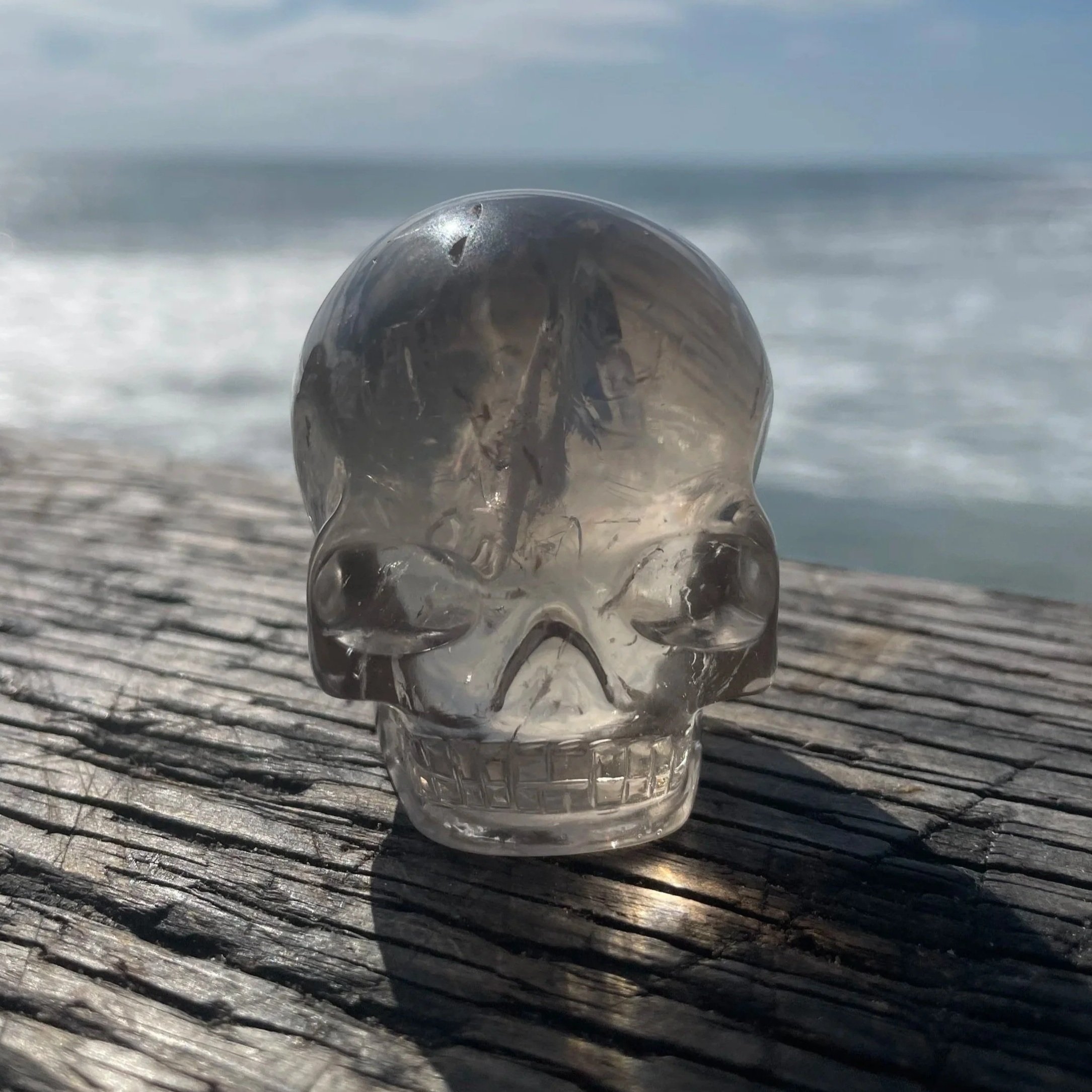 Close-up of a clear, transparent glass ball resting on a textured wooden surface, with ocean waves and cloudy sky in the background.
