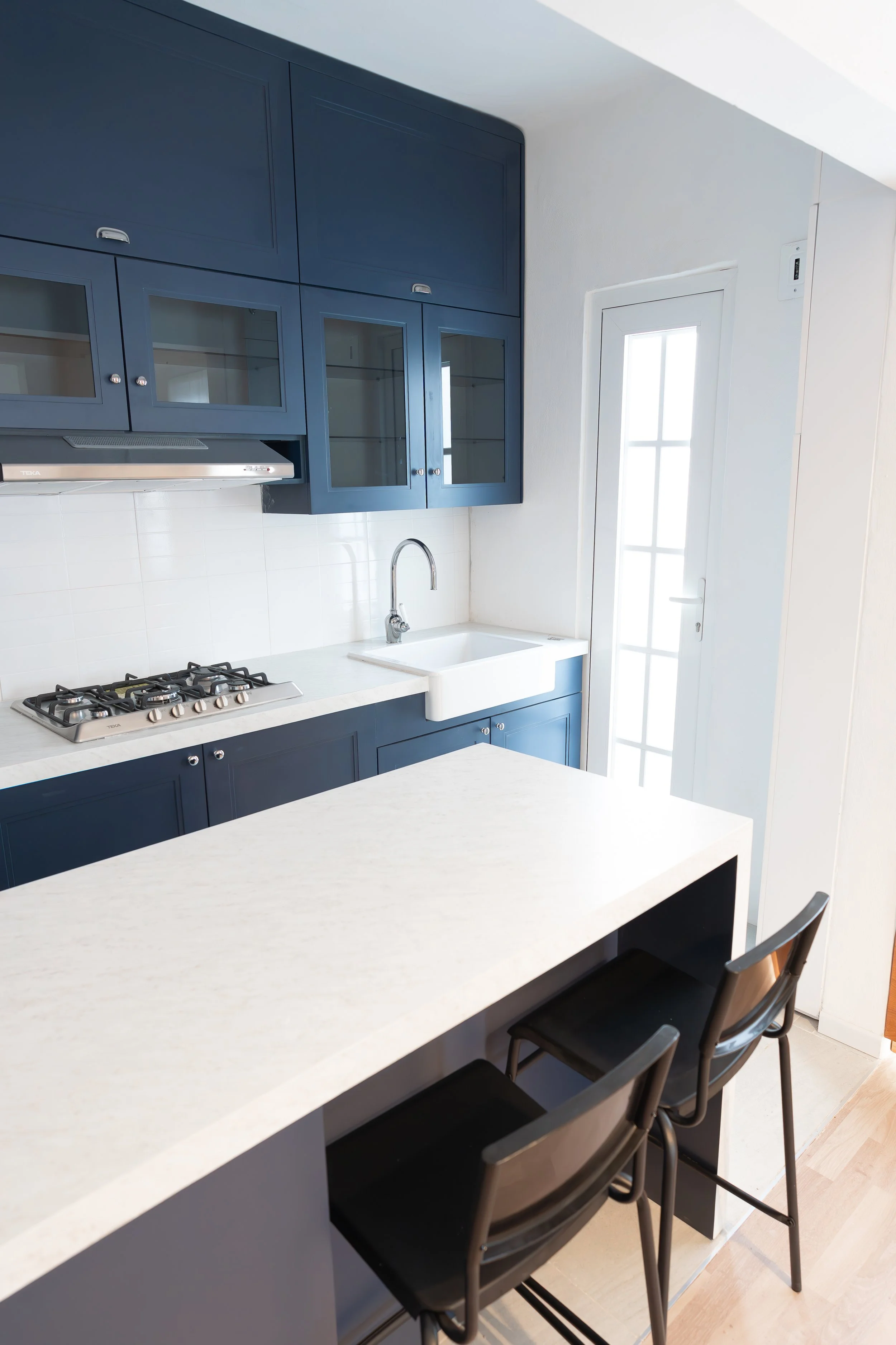 Cocina moderna con gabinetes azules, encimera de piedra blanca, estufa de dos quemadores, fregadero y sillas negras en una isla de cocina, puerta con ventana.