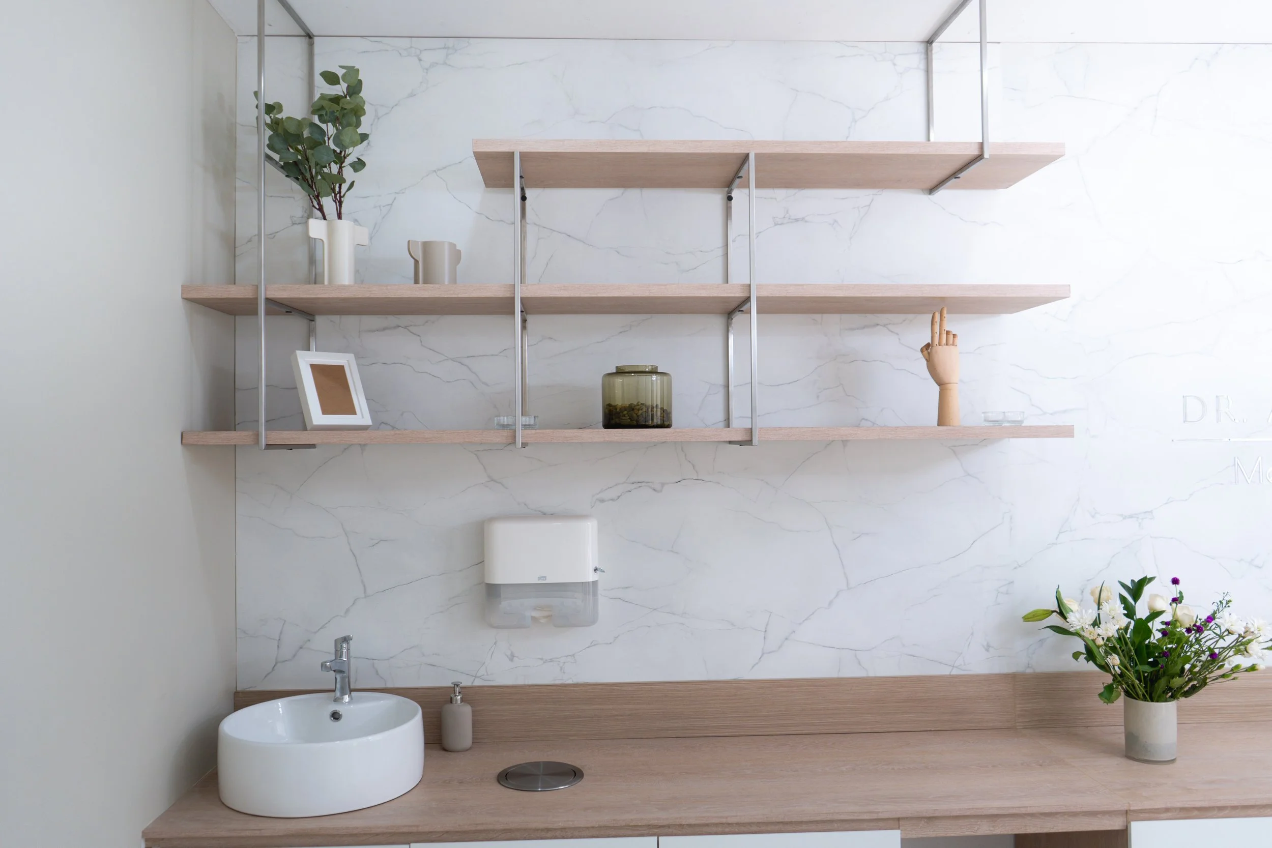 Estantería de madera y metal con plantas, marco, y objetos decorativos en pared de mármol. Planta con flores blancas en la esquina.