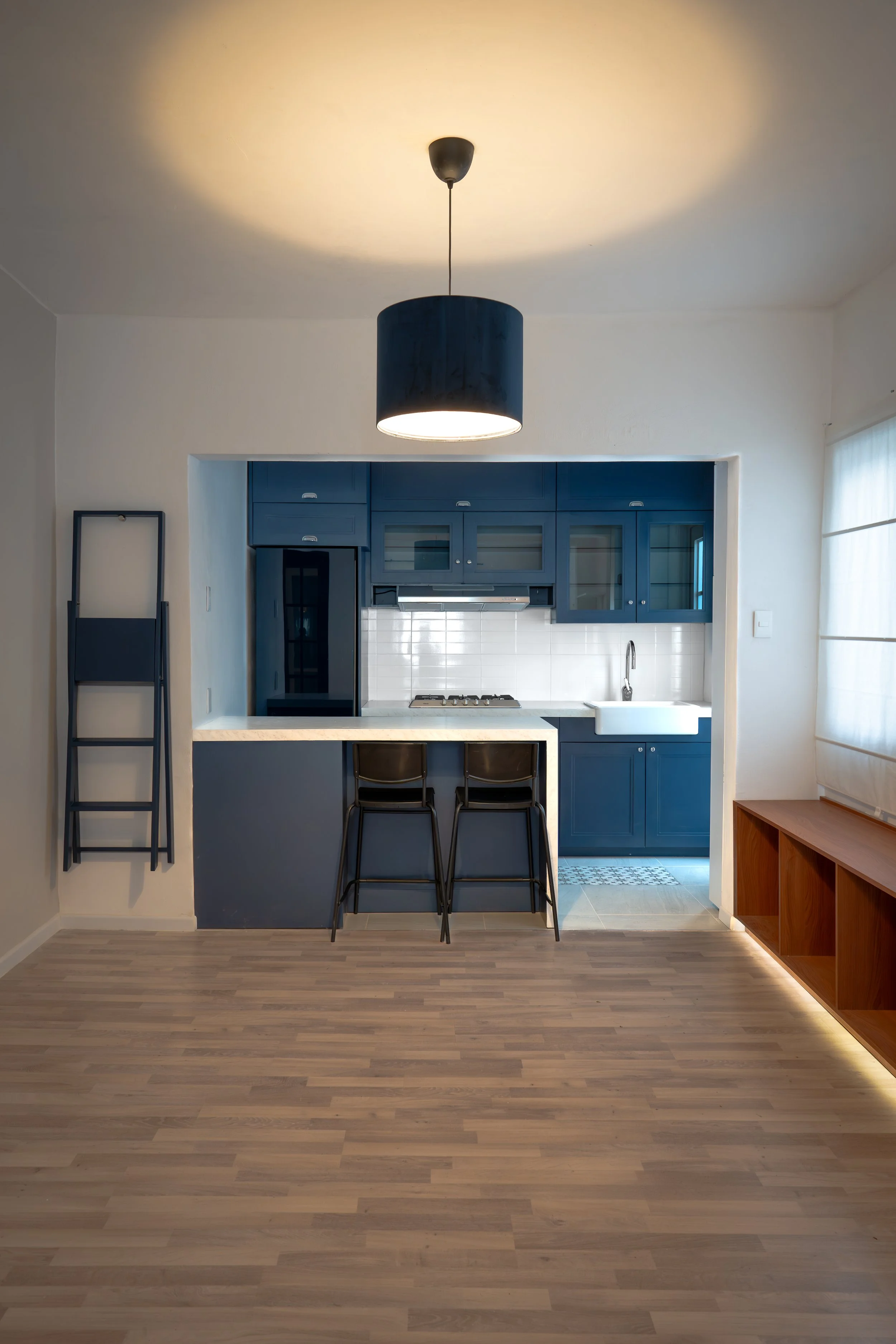 Cocina moderna con gabinetes azules, barra de desayuno con dos sillas negras, lámpara de techo grande y ventanas con cortinas blancas.
