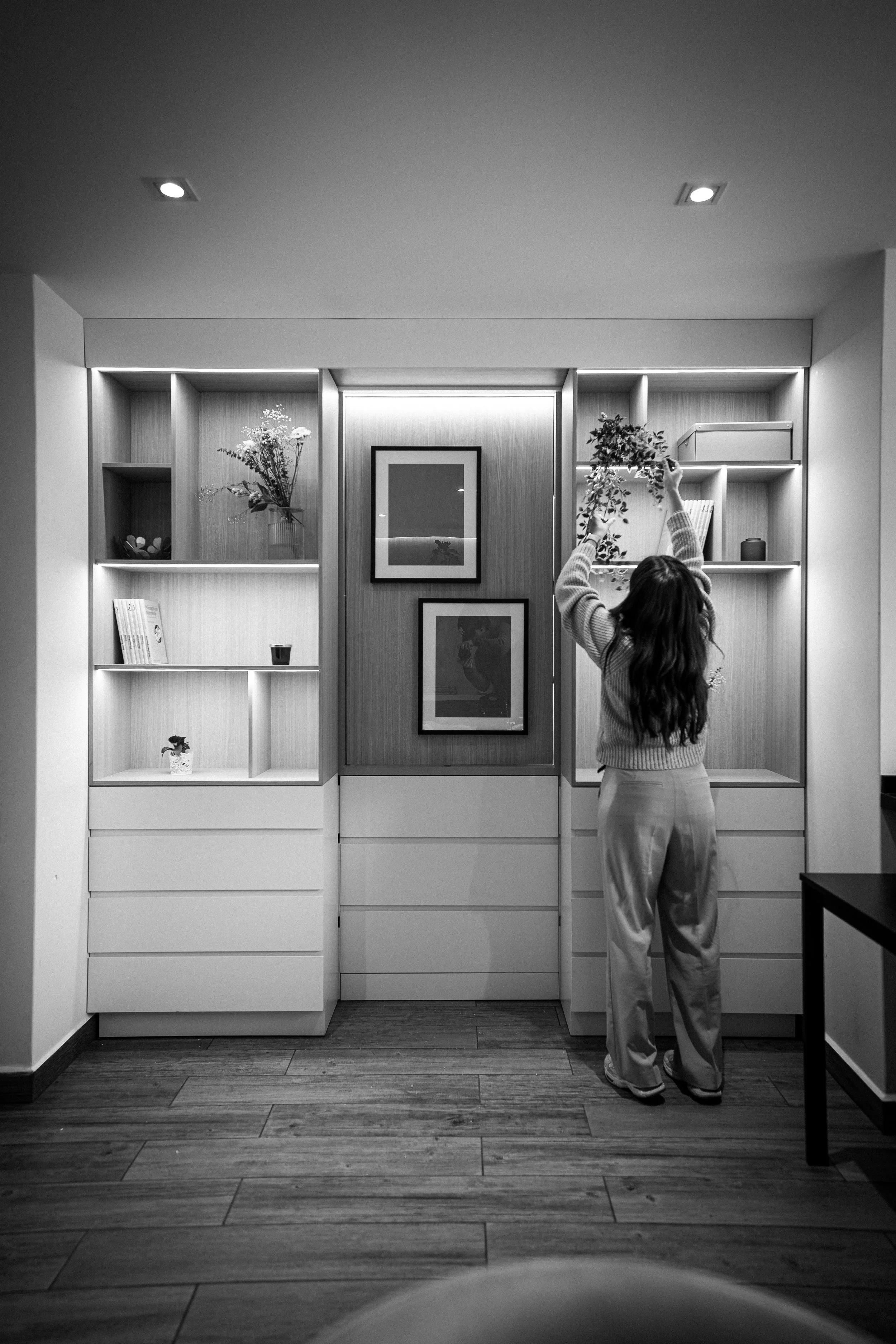 Una niña con pantalones claros y suéter largo, de espaldas, coloca flores en una repisa de madera en una pared con cuadros y estanterías con libros y decoraciones, todo en blanco y negro.