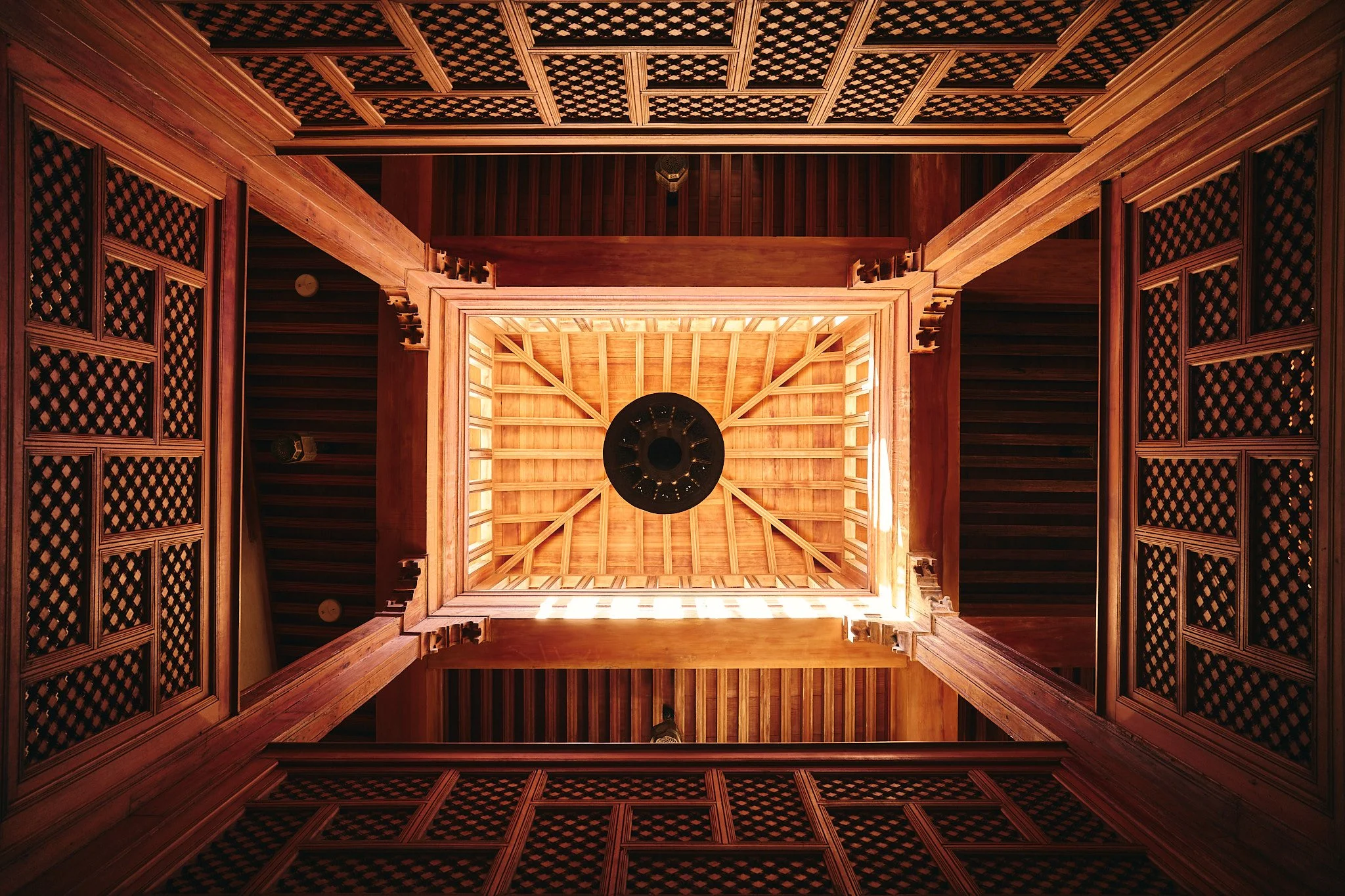 Perspectiva desde abajo hacia un techo de madera con detalles enrejados y un ventilador en el centro.