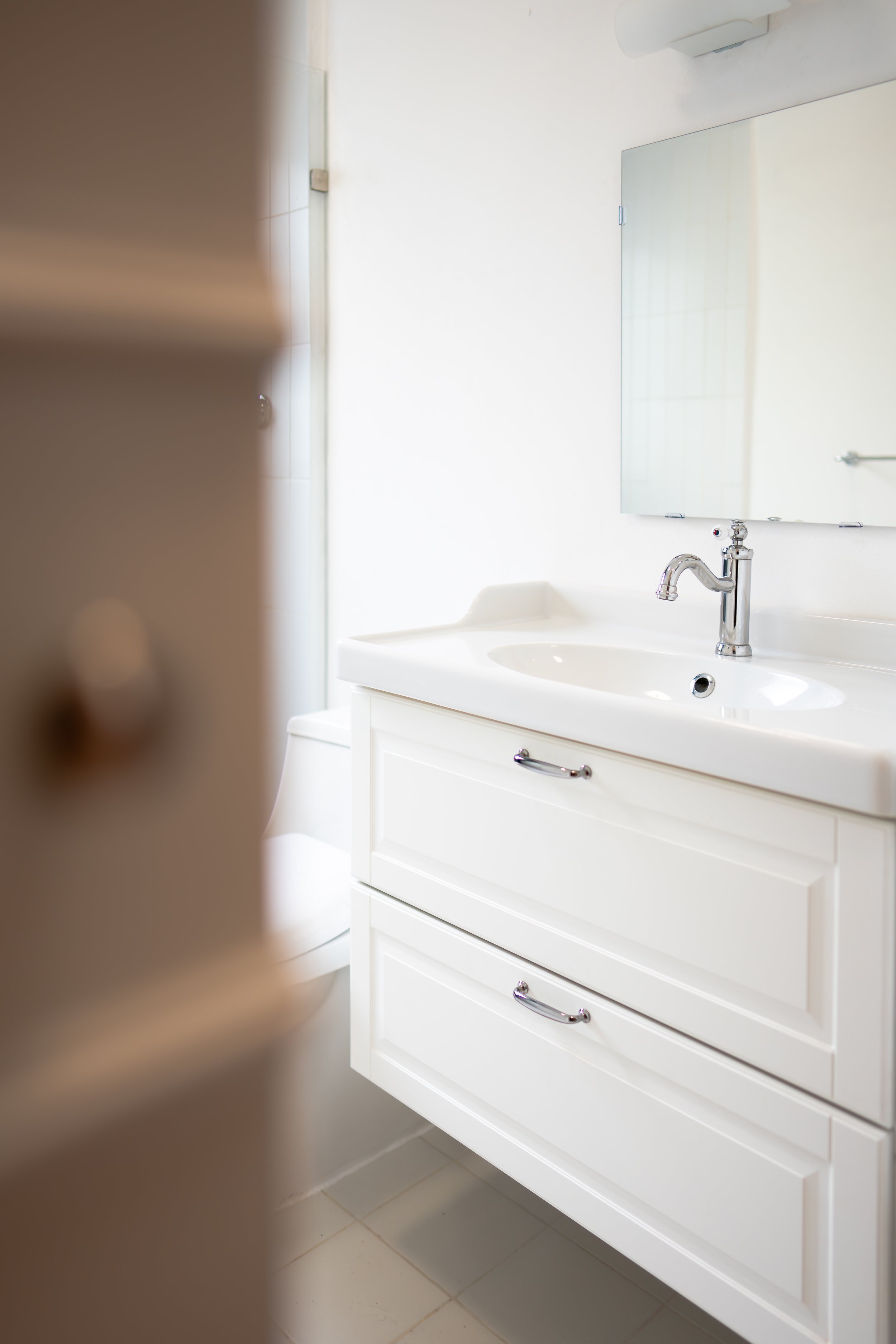 Espejo, lavabo y armario en un baño blanco, con una puerta y cerámica en el suelo.