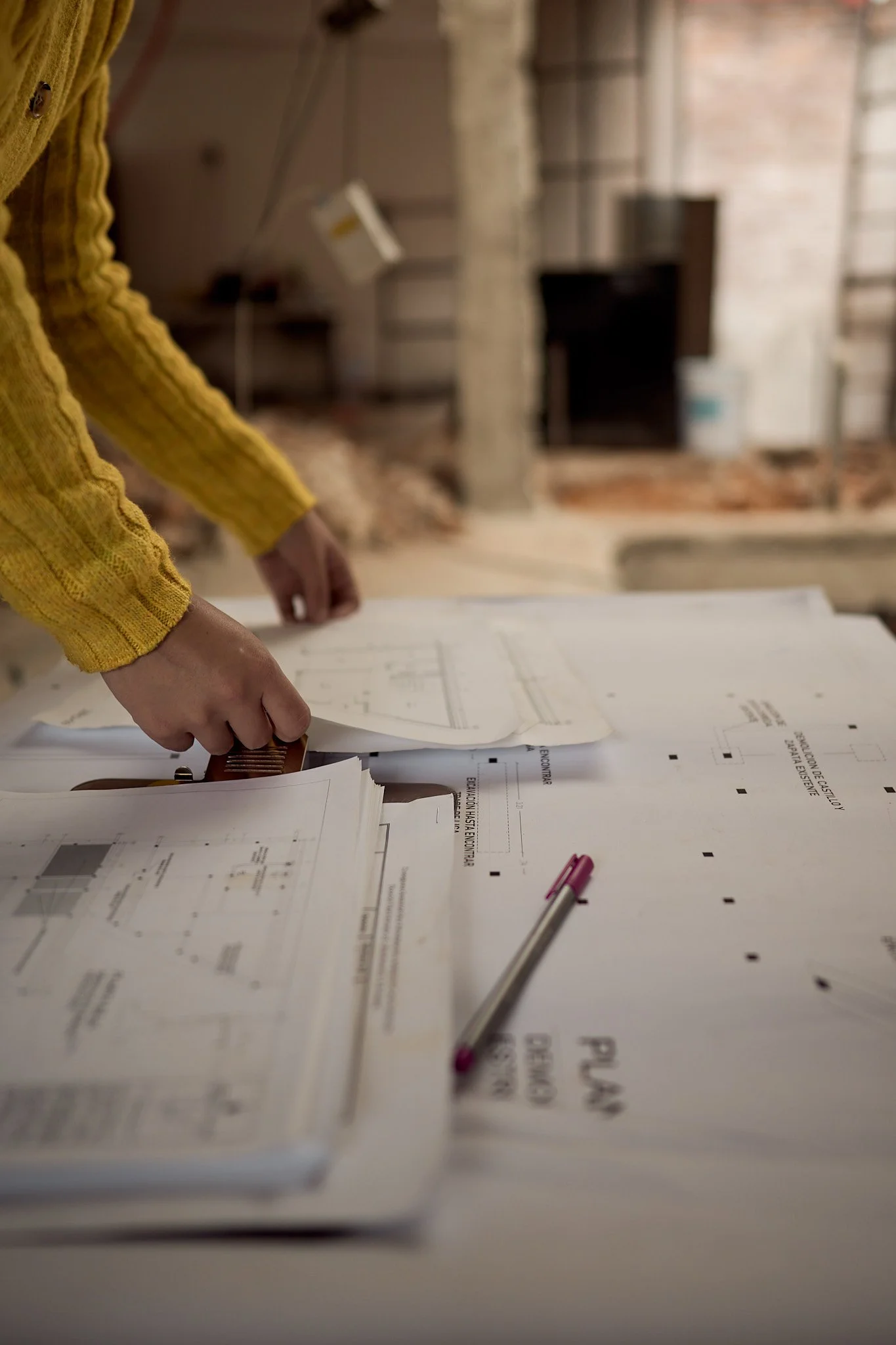 Persona revisando planos de construcción en una mesa con bolígrafo y documentos técnicos en un entorno de oficina o taller.