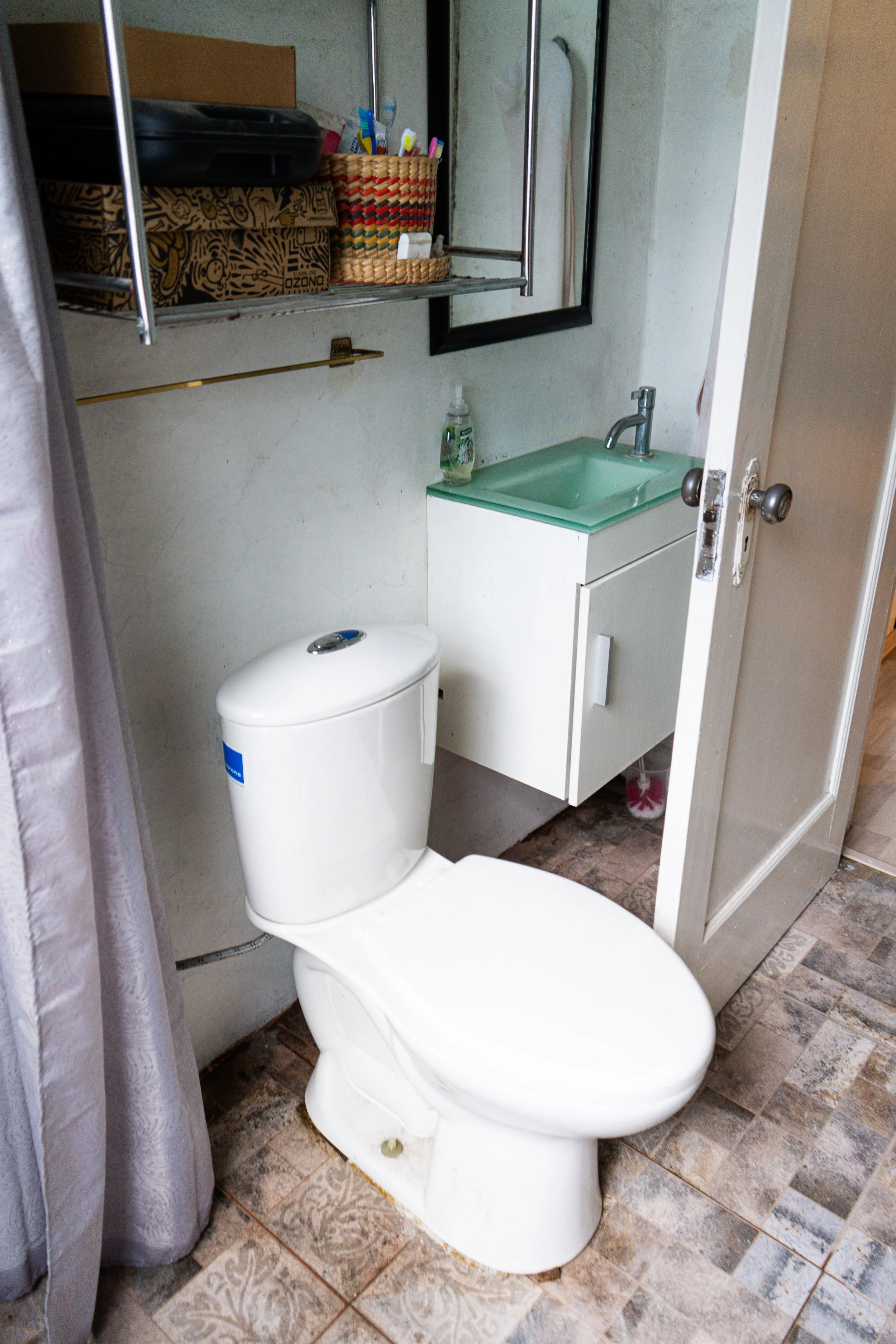 Baño con inodoro, lavabo pequeño con gabinete y espejo, estantería con artículos de higiene, y piso de baldosas decoradas.