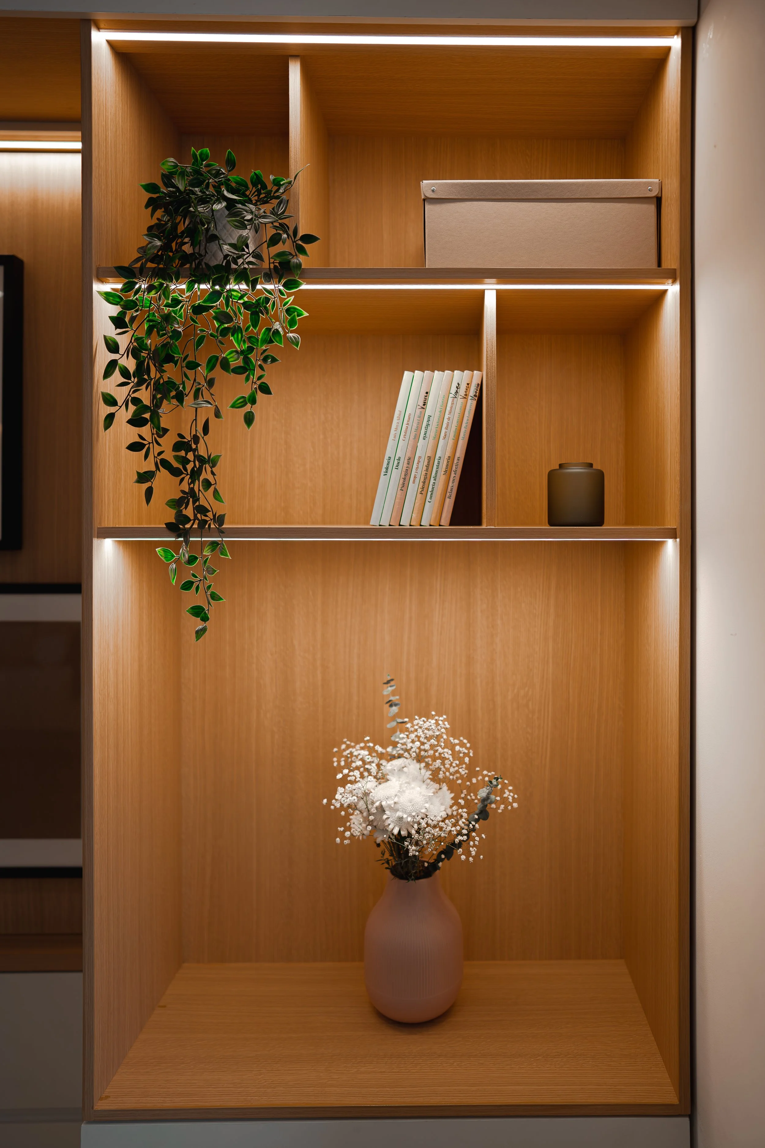 Estantería de madera con un libro, una caja, una planta colgante, una floristería con flores blancas en un jarrón y una lámpara LED en la parte superior, todo en un diseño moderno y minimalista.